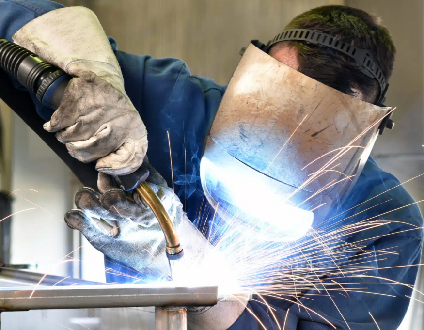 A welder in safety gear, including a face shield and gloves, welding metal with sparks flying.