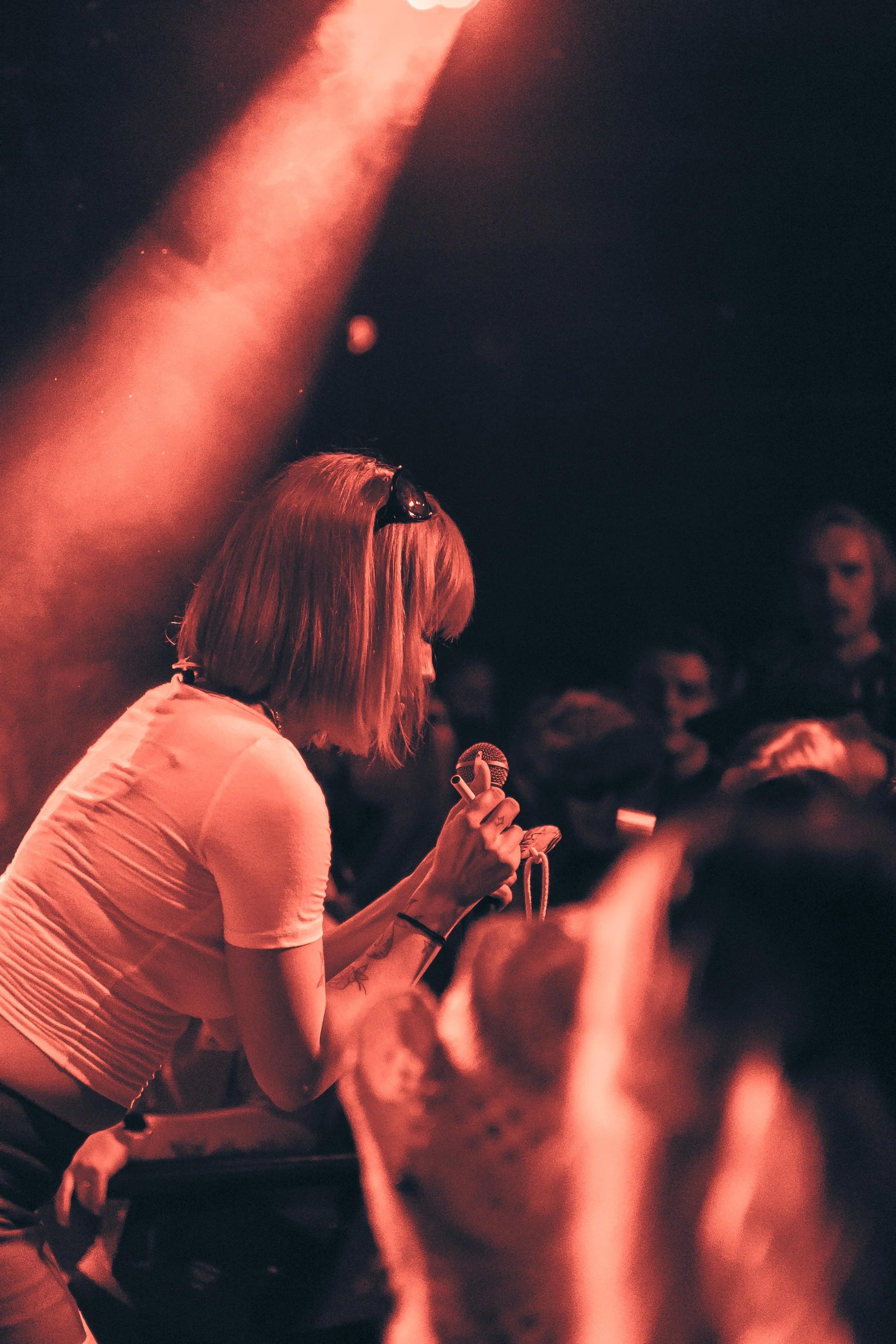 Nuori nainen laulaa mikrofonin kanssa keikalla pimeässä tilassa, jossa on muita ihmisiä ja kohtalainen valaistus.