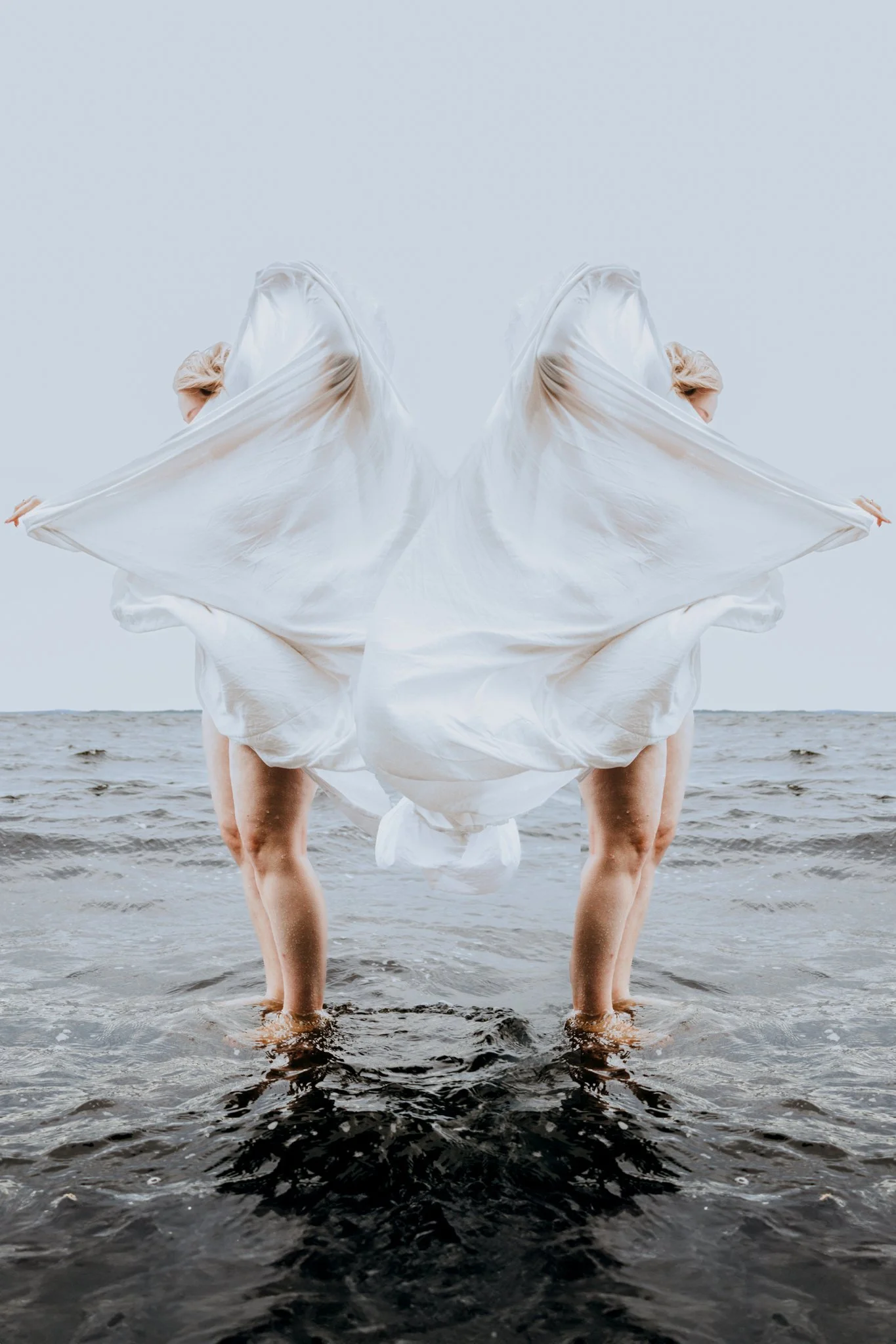 Kuvassa on kaksi ihmistä, jotka seisovat mereen päin ja ovat osittain peitetty valkoisella puvulla, jonka he pitävät levitettynä. Kuvan on symmetrinen ja lavastettu.