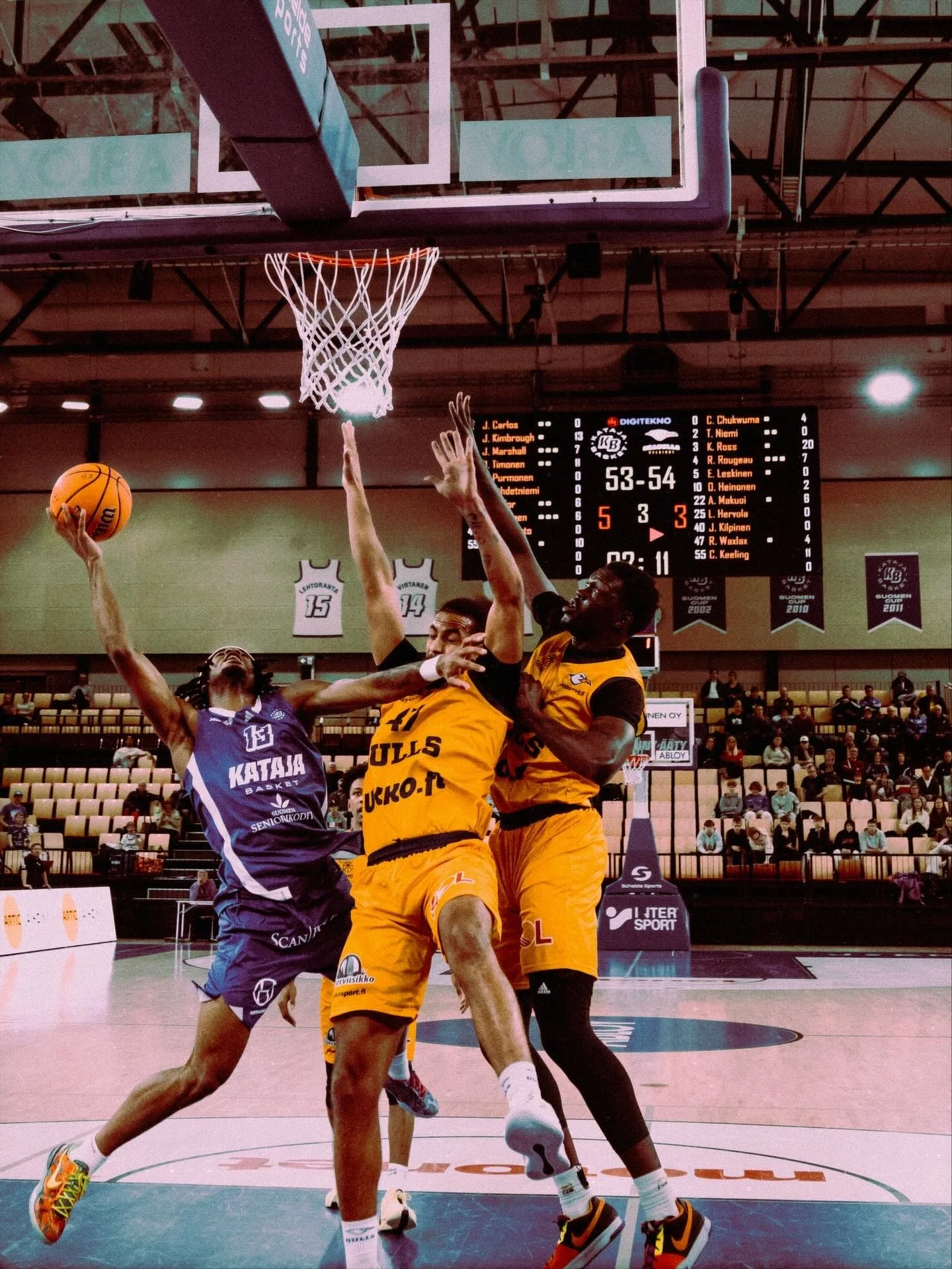 Koripallo-peli, kolme pelaajaa hyppää ilmaan yrittäen saada pallo koriin. Taustalla näkyy tulostaulu ja katsomo täynnä ihmisiä.
