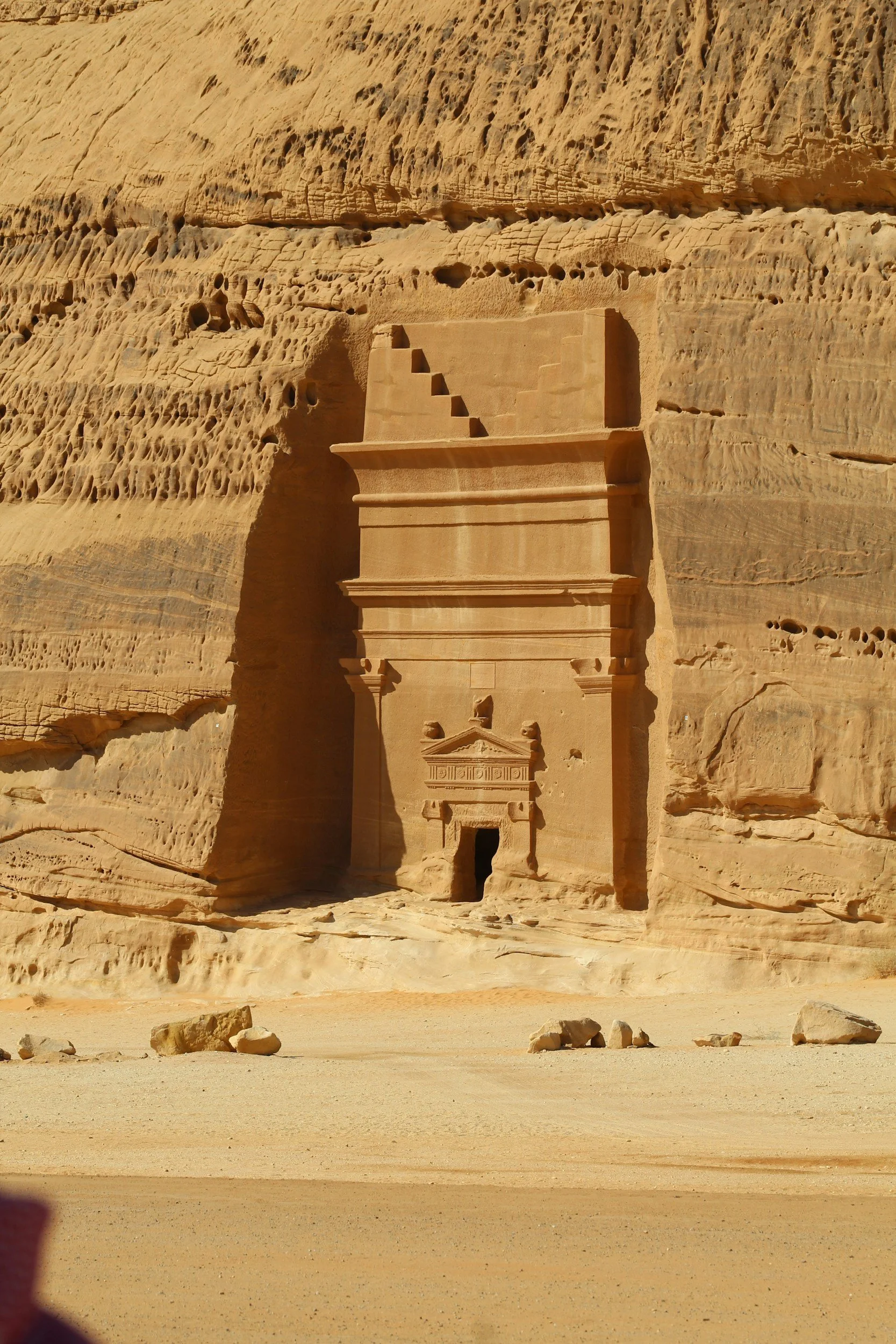 Ancient rock-cut tomb in Saudi Arabia carved into a sandstone cliff with detailed façade and stairs, located in a desert environment.