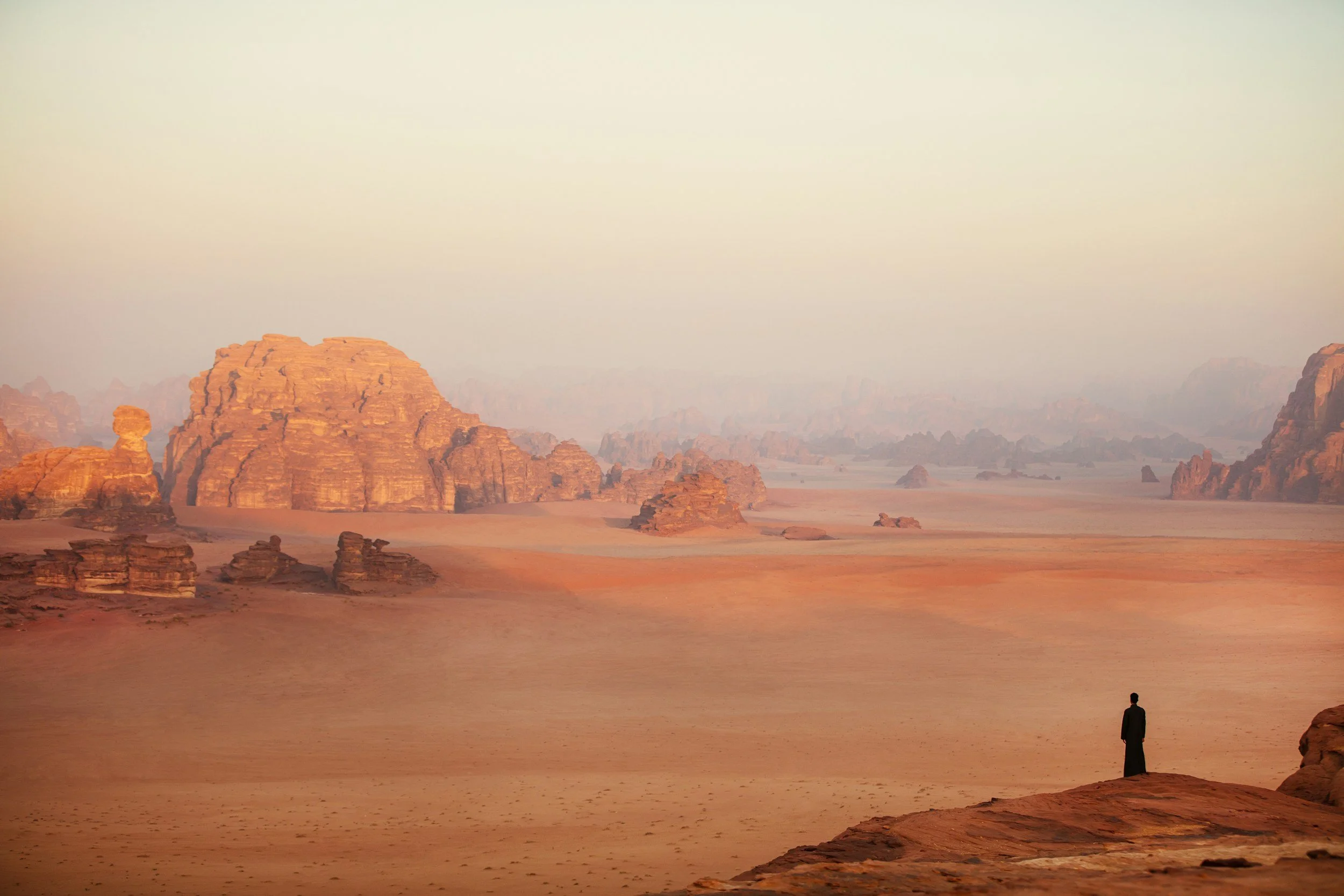 A solitary person dressed in black standing on a desert rock formation, overlooking a vast, rocky desert landscape with large sandstone formations in the distance, under a hazy sky.