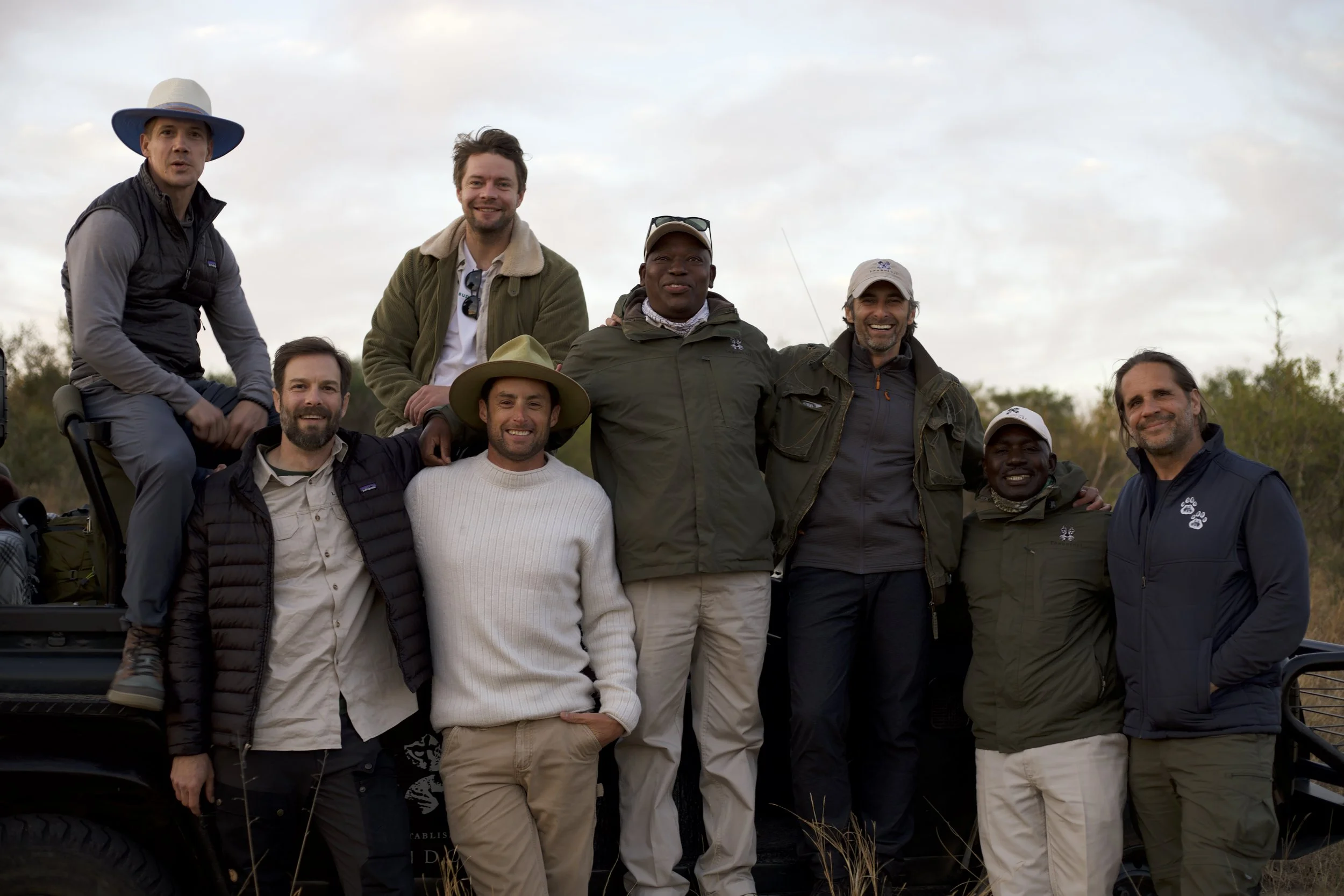 A group of nine men posing outdoors during daytime, some standing, some sitting on a vehicle, with a natural landscape background.
