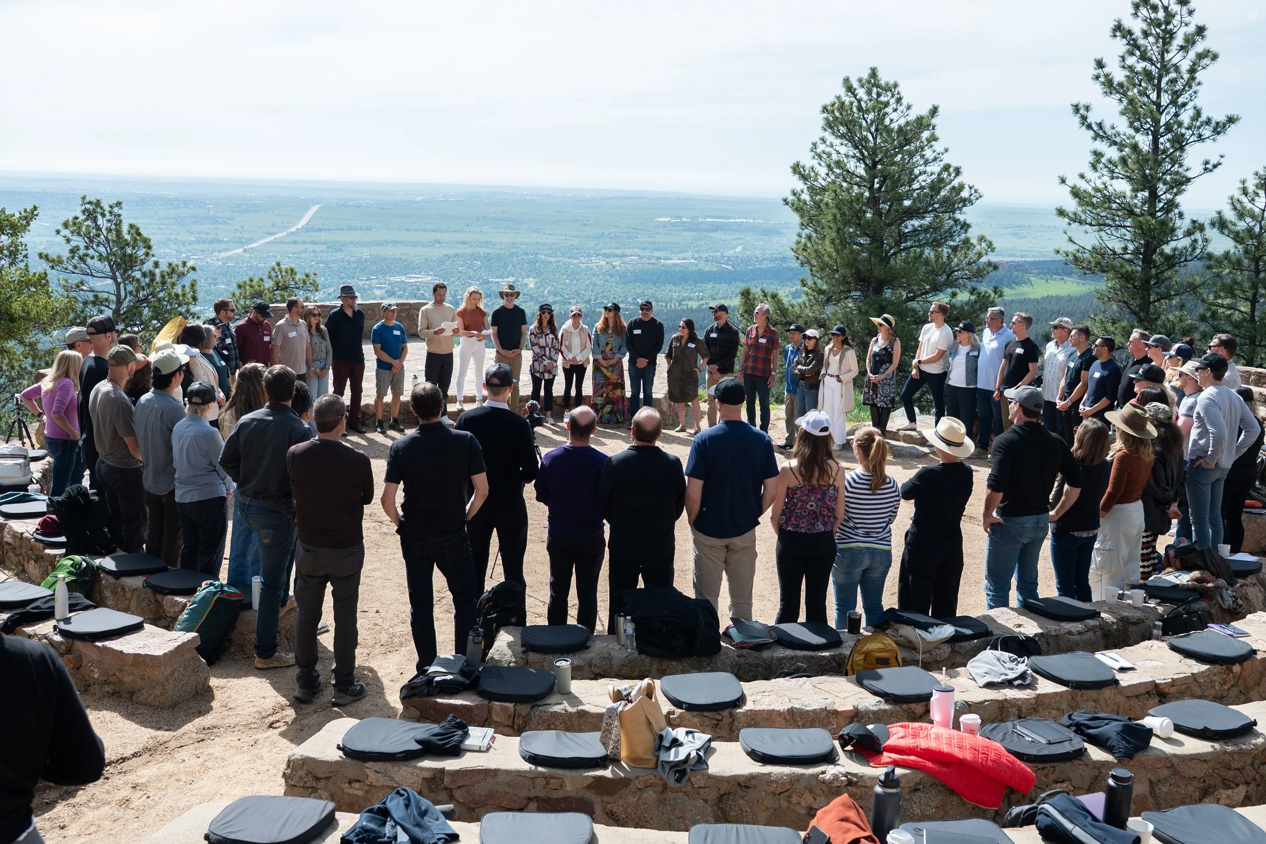 Large group of people gathered outdoors in a semi-circle, listening to a speaker at an outdoor event with a scenic view of trees and distant landscape in the background, some sitting on stone benches with belongings around.