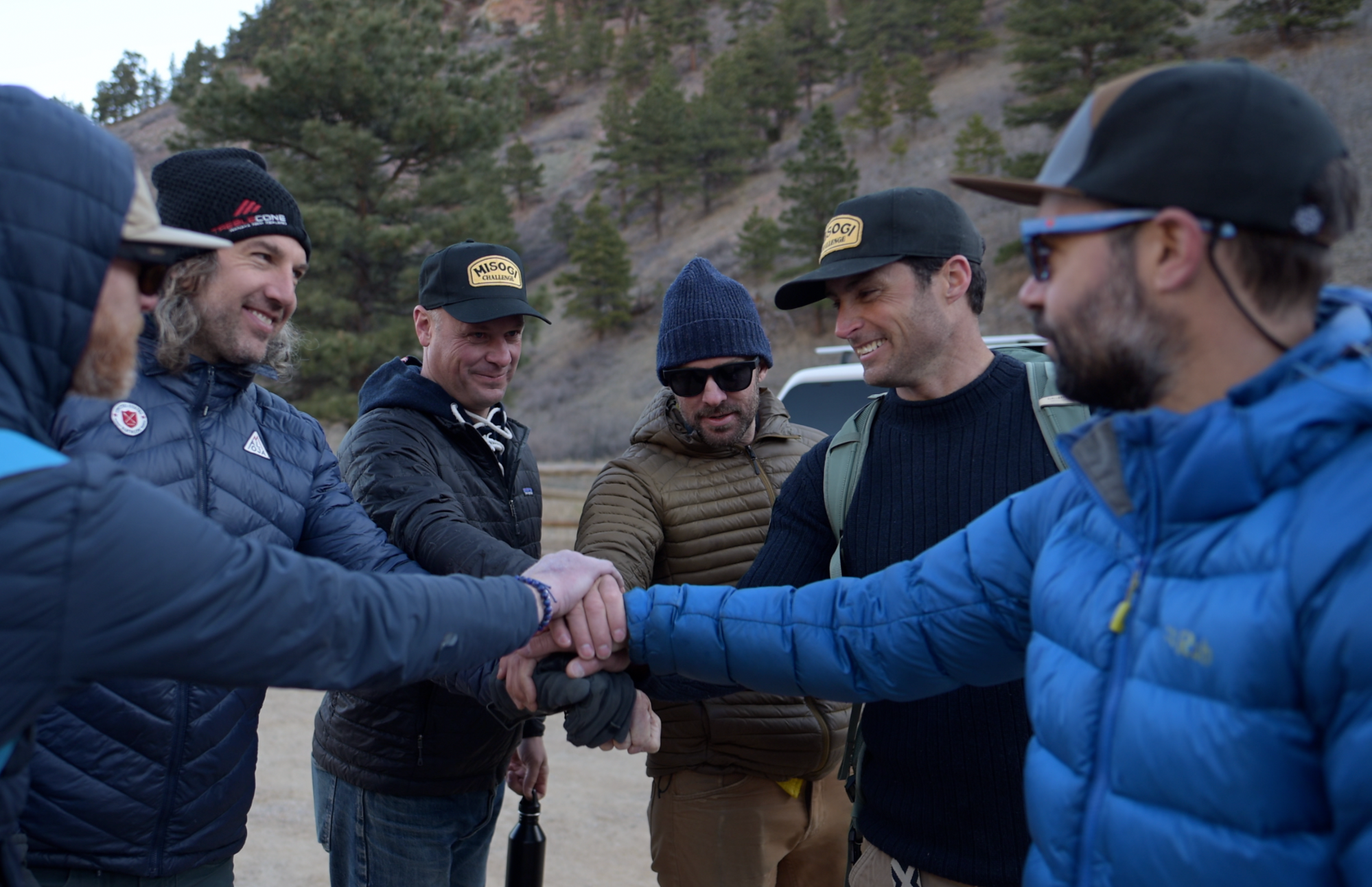 Group of men outdoors in a mountainous area, shaking hands and smiling, wearing jackets and caps.