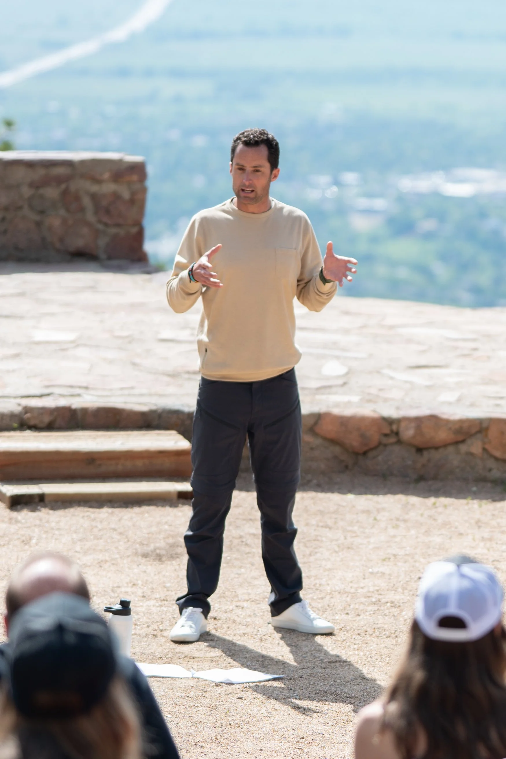 A man is giving a talk or presentation outdoors, with a scenic mountainous background, to an audience seated on the ground.