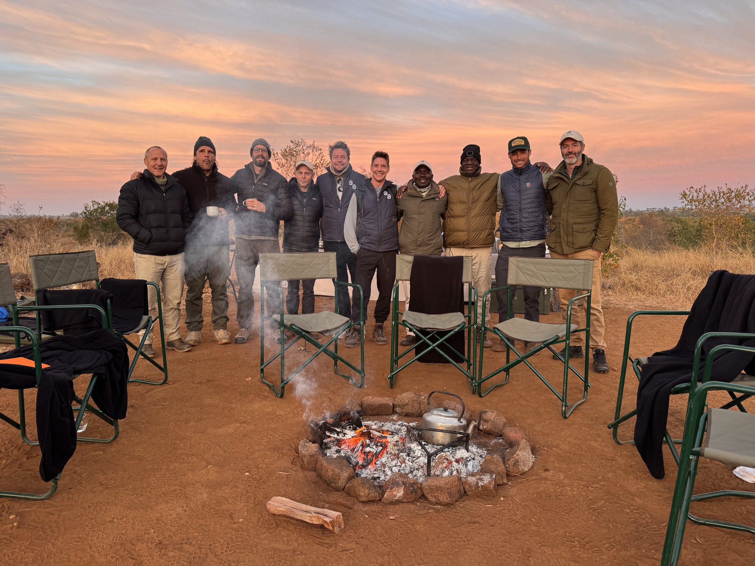 Group of eleven people standing outdoors around a campfire at sunset, with chairs and a kettle on the fire, in a natural, open landscape.
