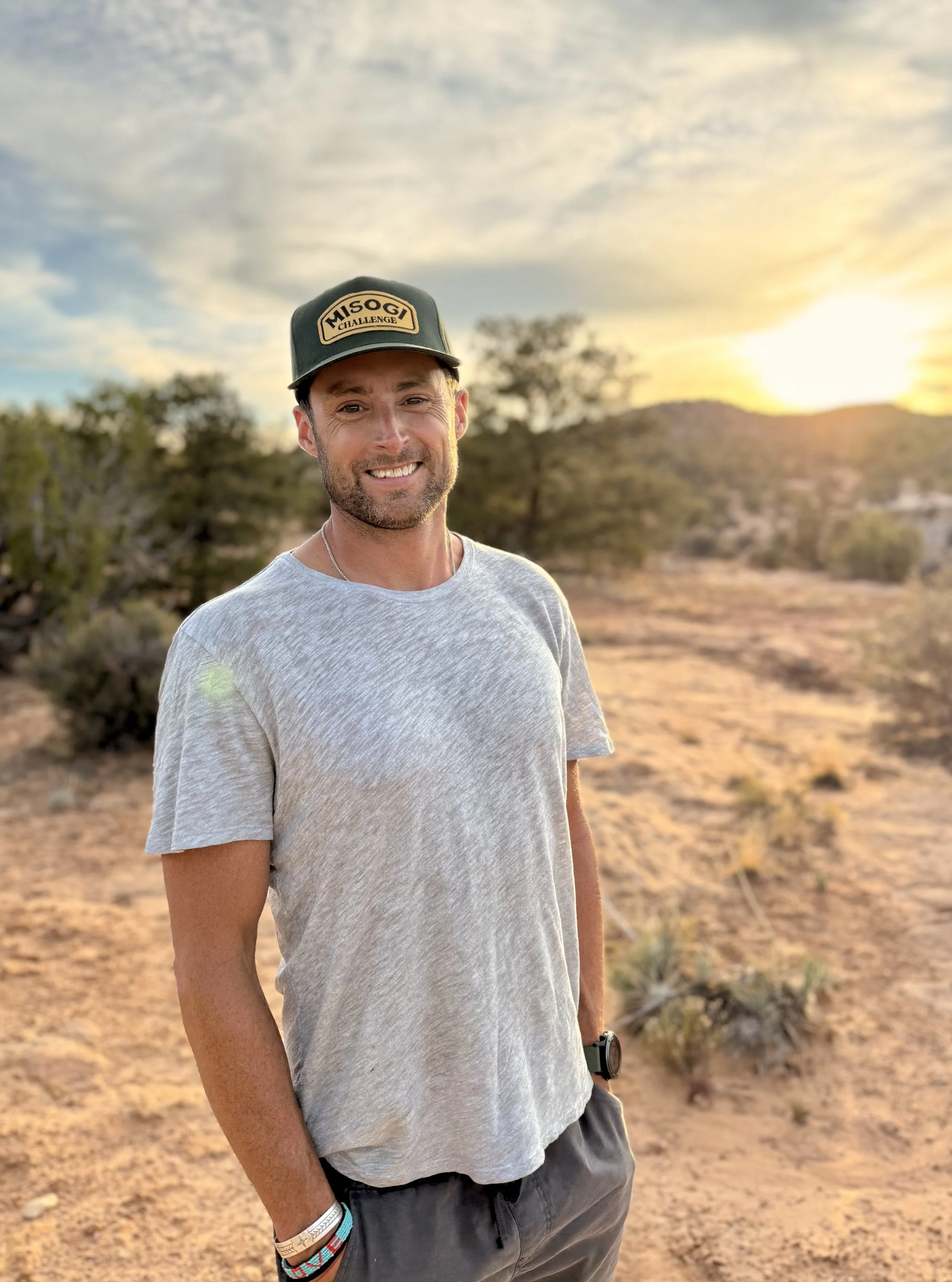A man smiling outdoors at sunset in a desert landscape with sparse bushes and mountains in the background, wearing a gray t-shirt, a black cap with a patch, and accessories on his wrist.