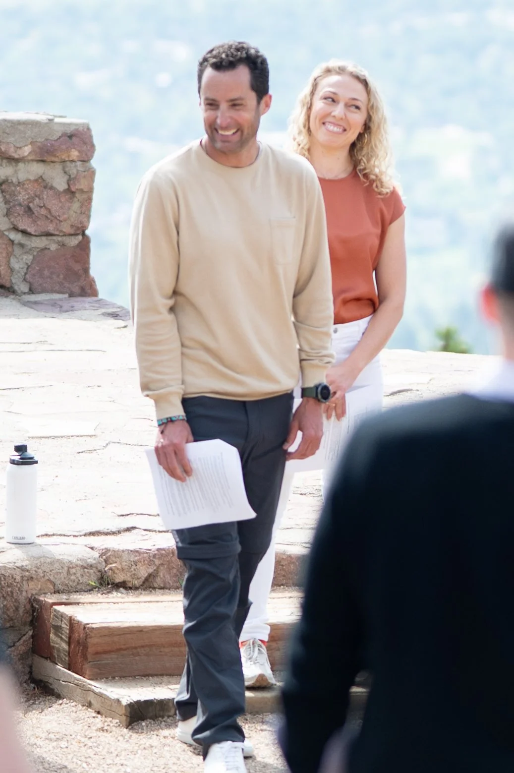 A man and woman standing outdoors, smiling during what appears to be a wedding or celebration, with a scenic mountain background.