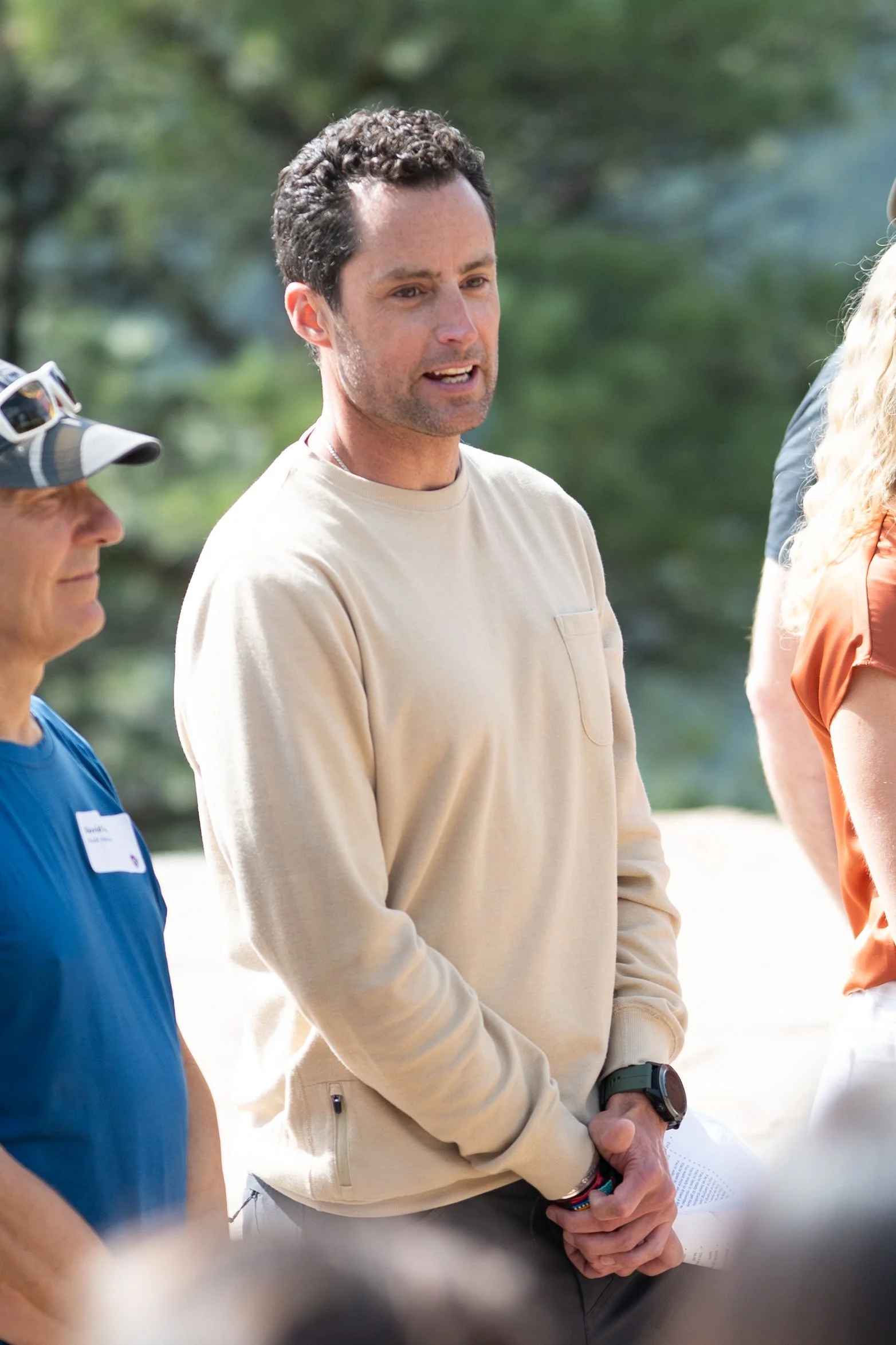 A man with dark, curly hair, wearing a beige long-sleeve shirt and a smartwatch, speaking to people outdoors, with a blurred background of green trees.