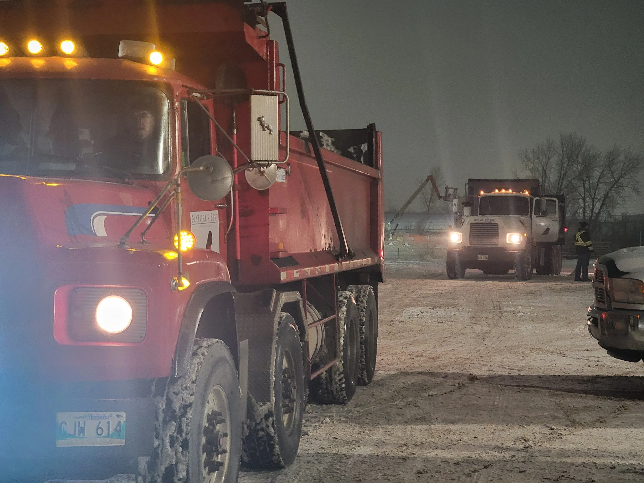 Two large red trucks with snow-covered tires at an outdoor site during winter night, one on the left with a partially visible license plate from Manitoba and one in the background with pavement hose attached, with a person in safety gear standing near the right truck and a leafless tree behind.