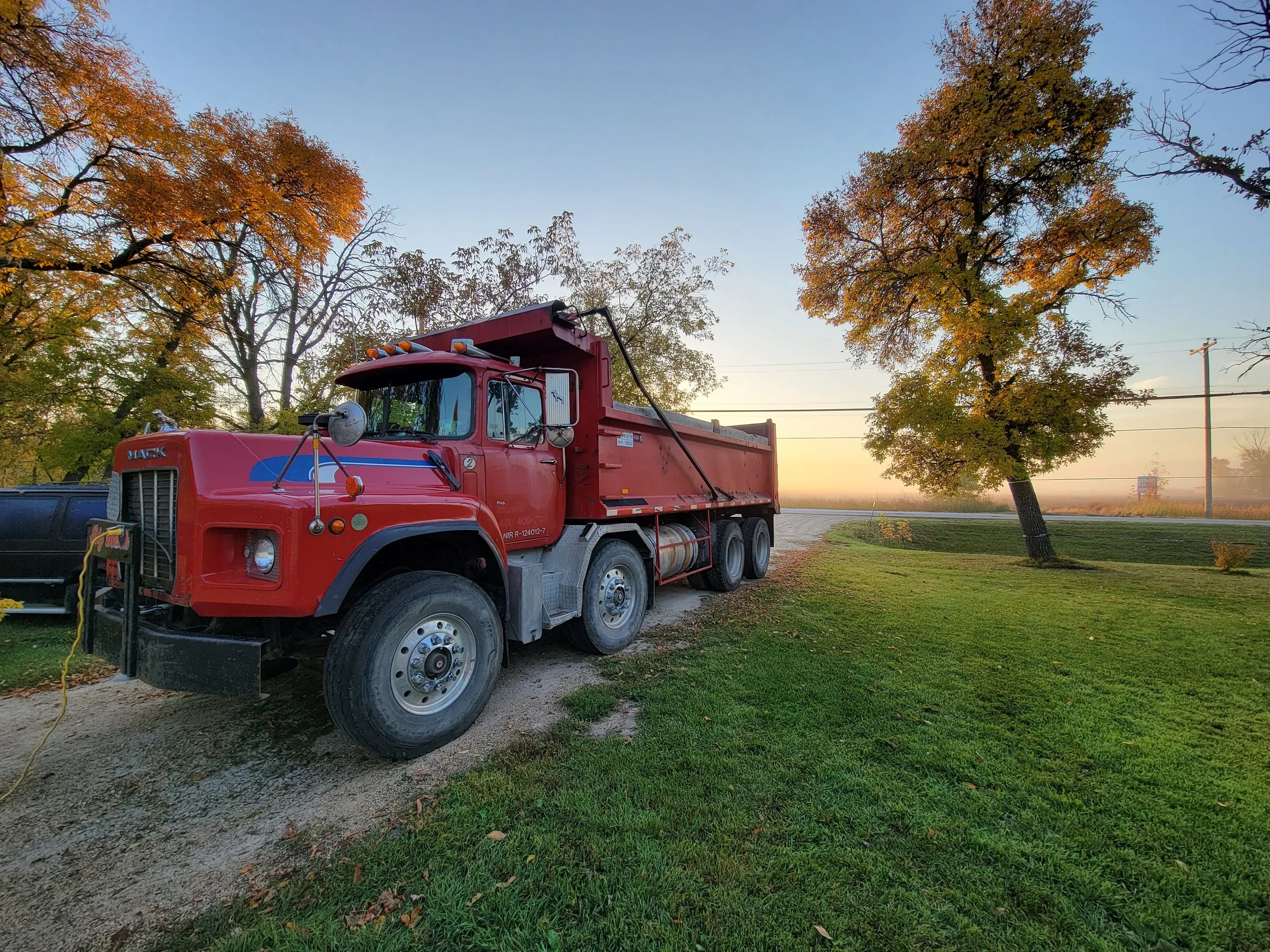 Red dump truck parked on a grassy area next to a dirt road with trees showing fall foliage in the background at sunset.