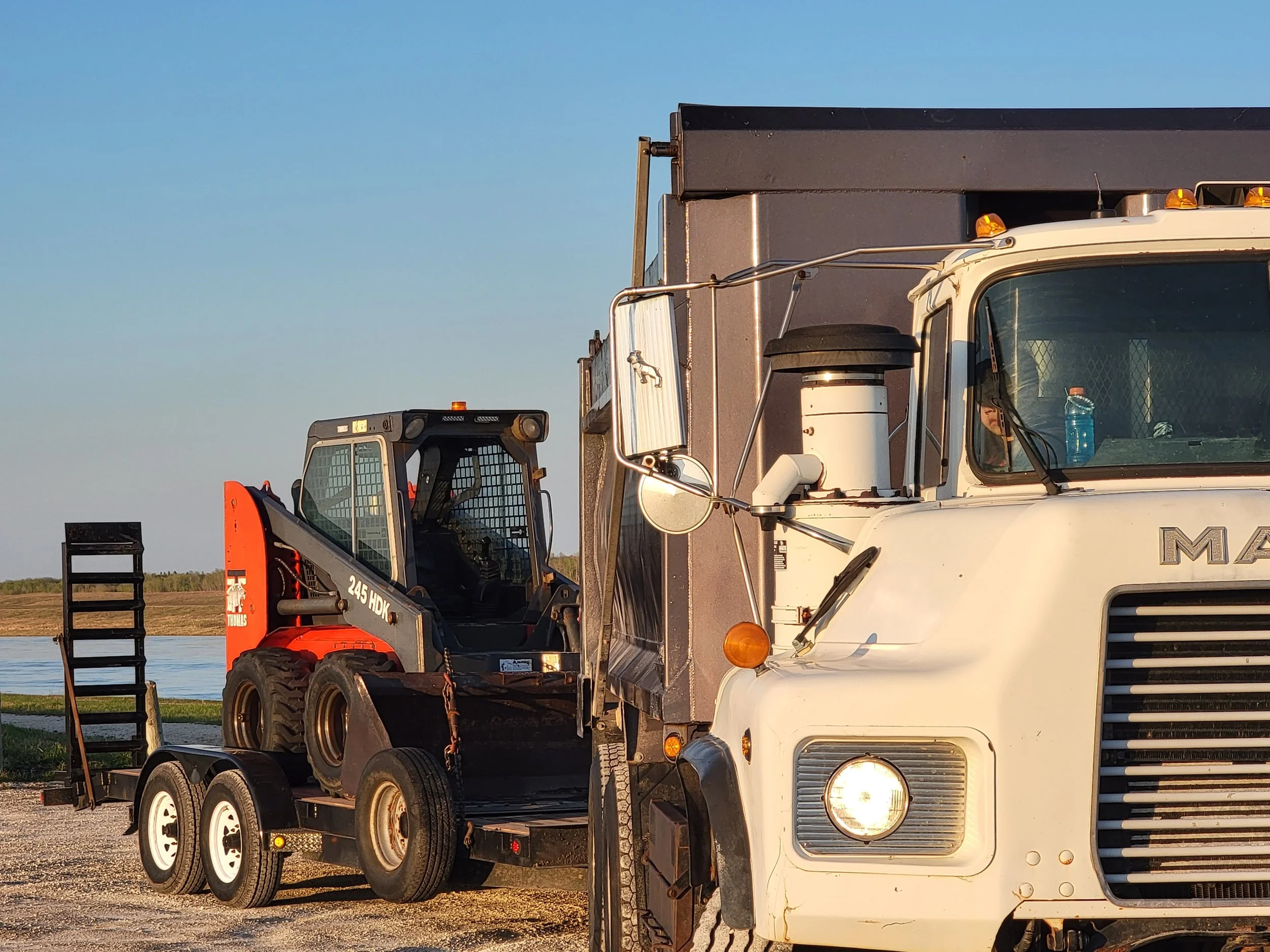 White tow truck hauling a skid steer loader on a trailer, near a lake, in the late afternoon.