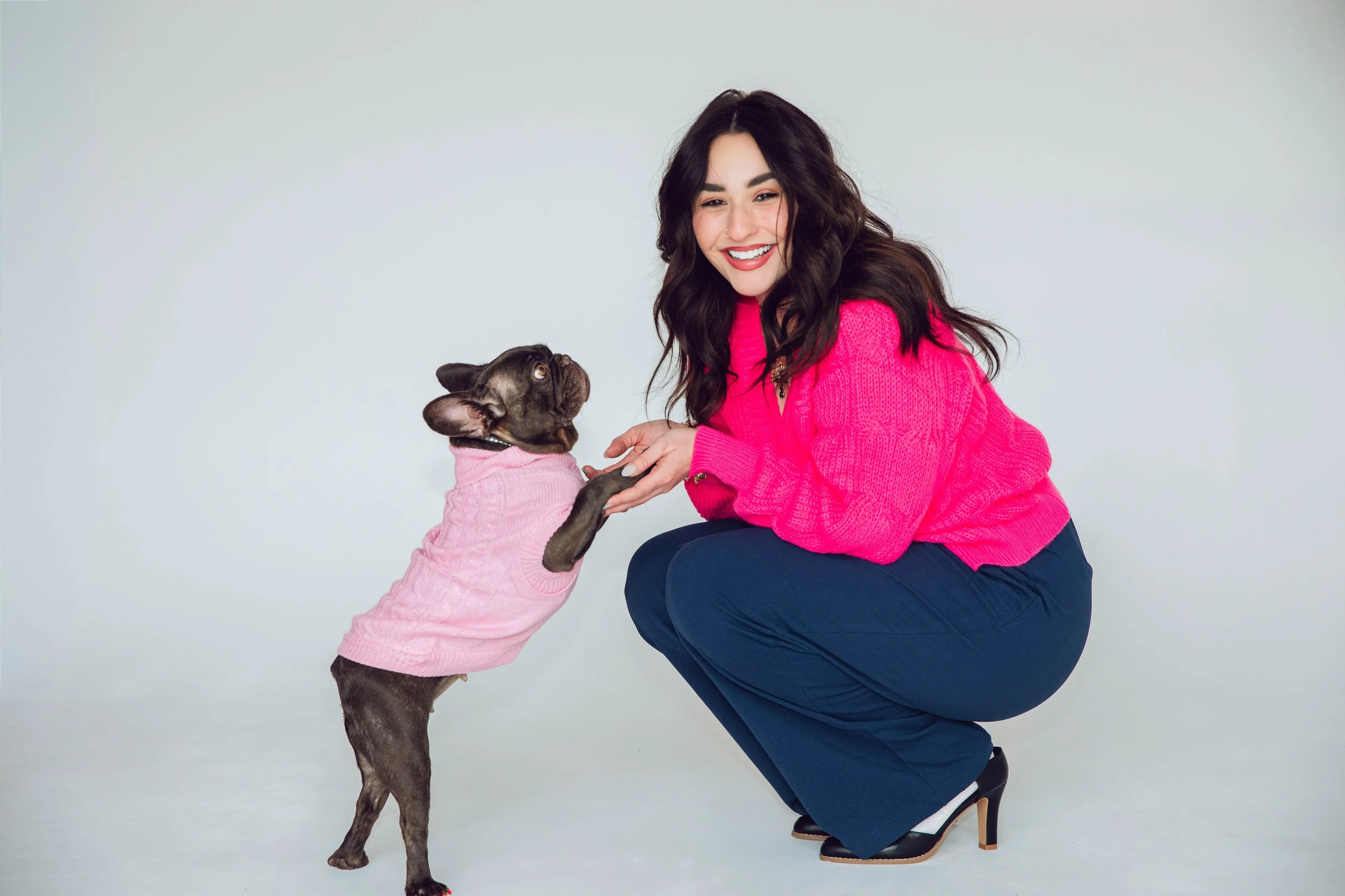 A woman in a pink sweater and navy blue pants crouches and smiles at a small dog wearing a pink sweater, with their paws clasped together.