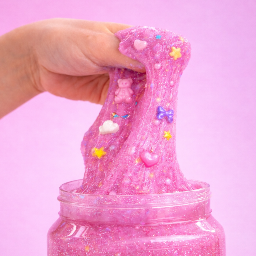 Pink slime with glitter, decorated with small candies including a teddy bear, hearts, stars, and bows, being pulled out of a jar against a pink background.