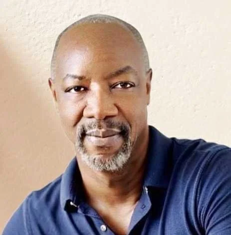 Close-up of a middle-aged man with a mustache and goatee, wearing a dark blue collared shirt, standing against a light-colored wall.