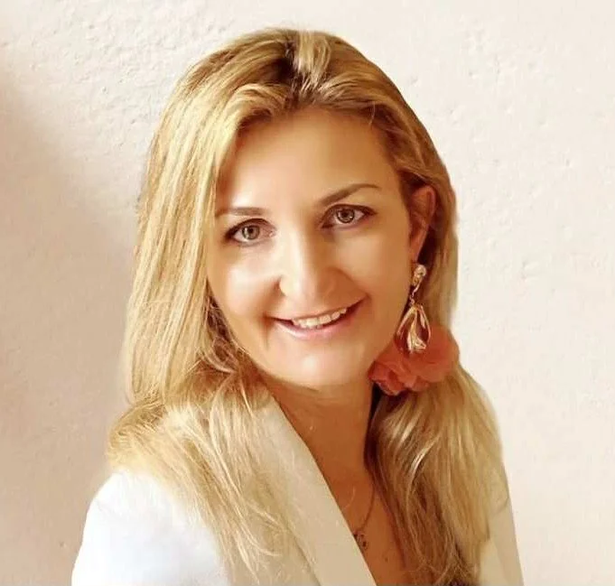 A woman with blonde hair and a warm smile, wearing a white top and a statement earring with a pink flower, standing against a plain light-colored wall.