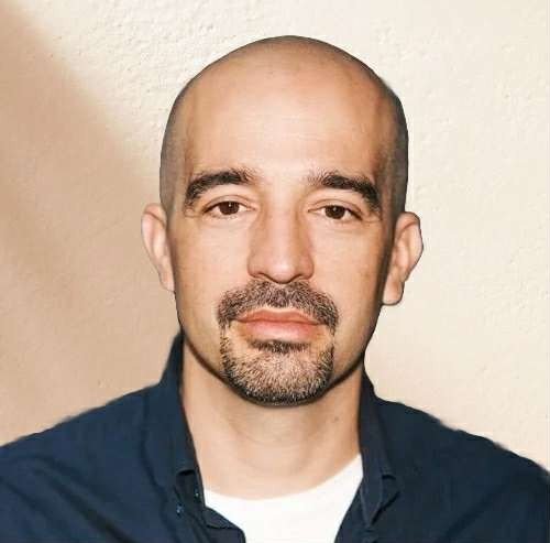 Headshot of a bald man with a short beard, wearing a dark jacket and white shirt, against a plain beige background.