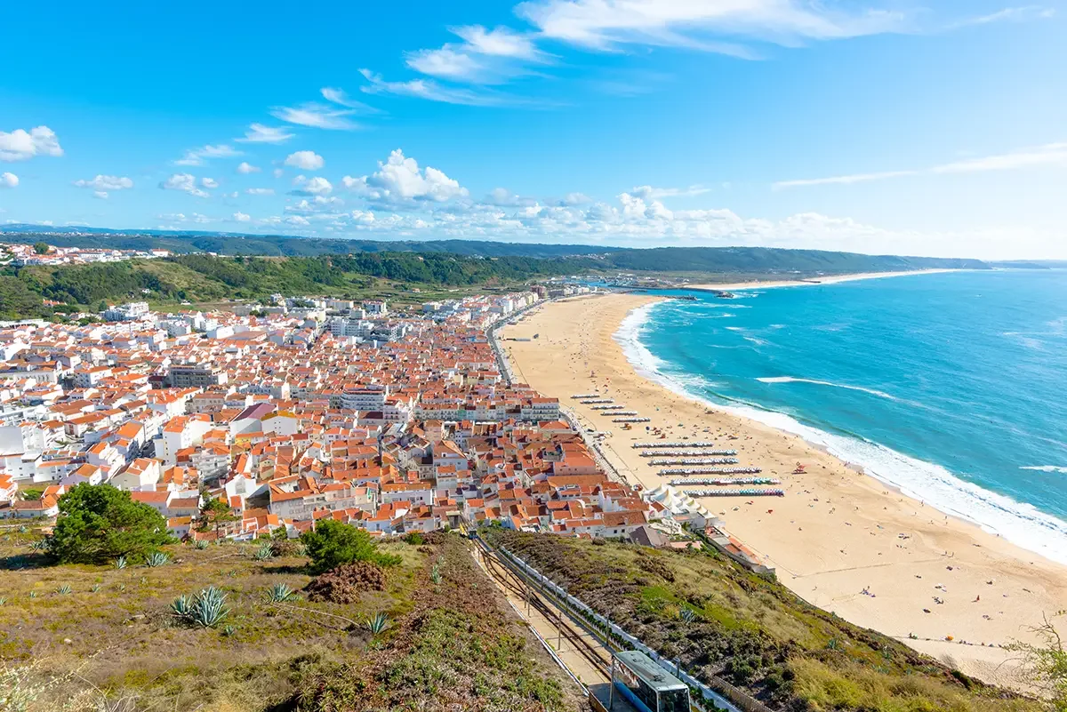 A coastal town with a sandy beach, colorful rooftops, and a train approaching from a hillside.