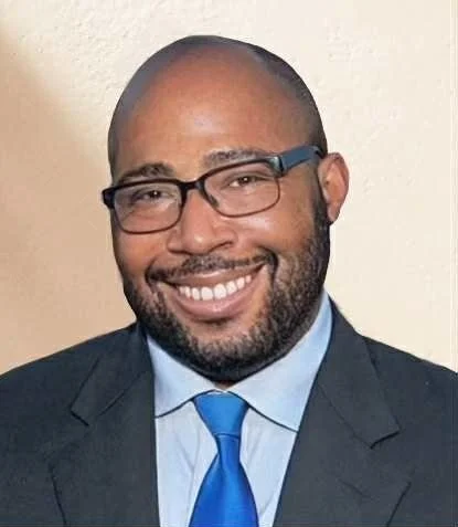 Smiling man in a suit with glasses, wearing a light blue shirt and blue tie, against a neutral background.