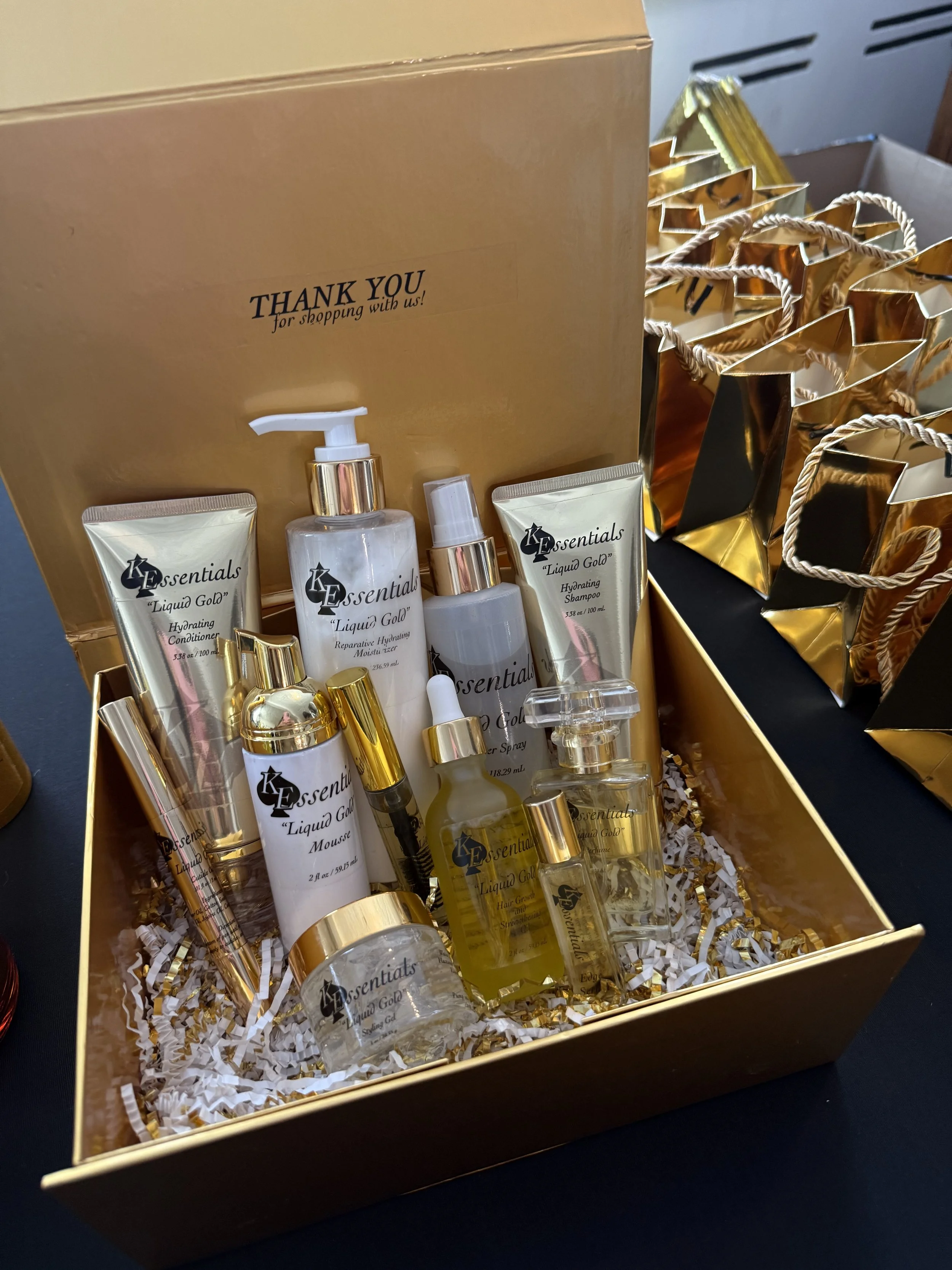 A gold-colored gift box filled with assorted beauty products and skincare items. The products include tubes and bottles labeled 'K Essentials Liquid Gold' and come with gold packaging. The box is decorated with shredded paper for cushioning.