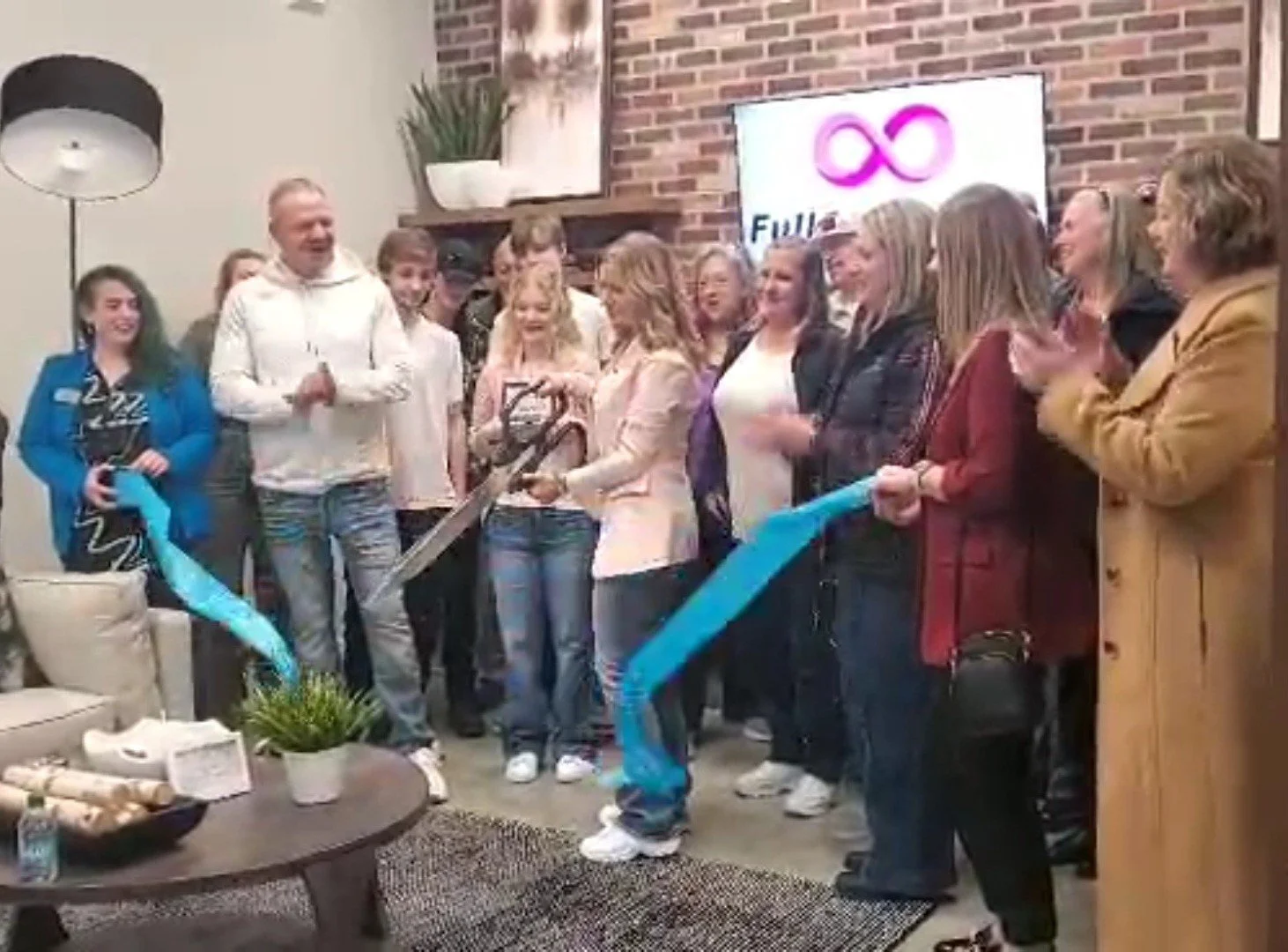 People gathered indoors at a celebration, some clapping and smiling, with a woman cutting a ribbon. A television screen with a logo and the word 'FULL' is visible in the background, along with brick and painted walls, a table with decor, and a tall floor lamp.