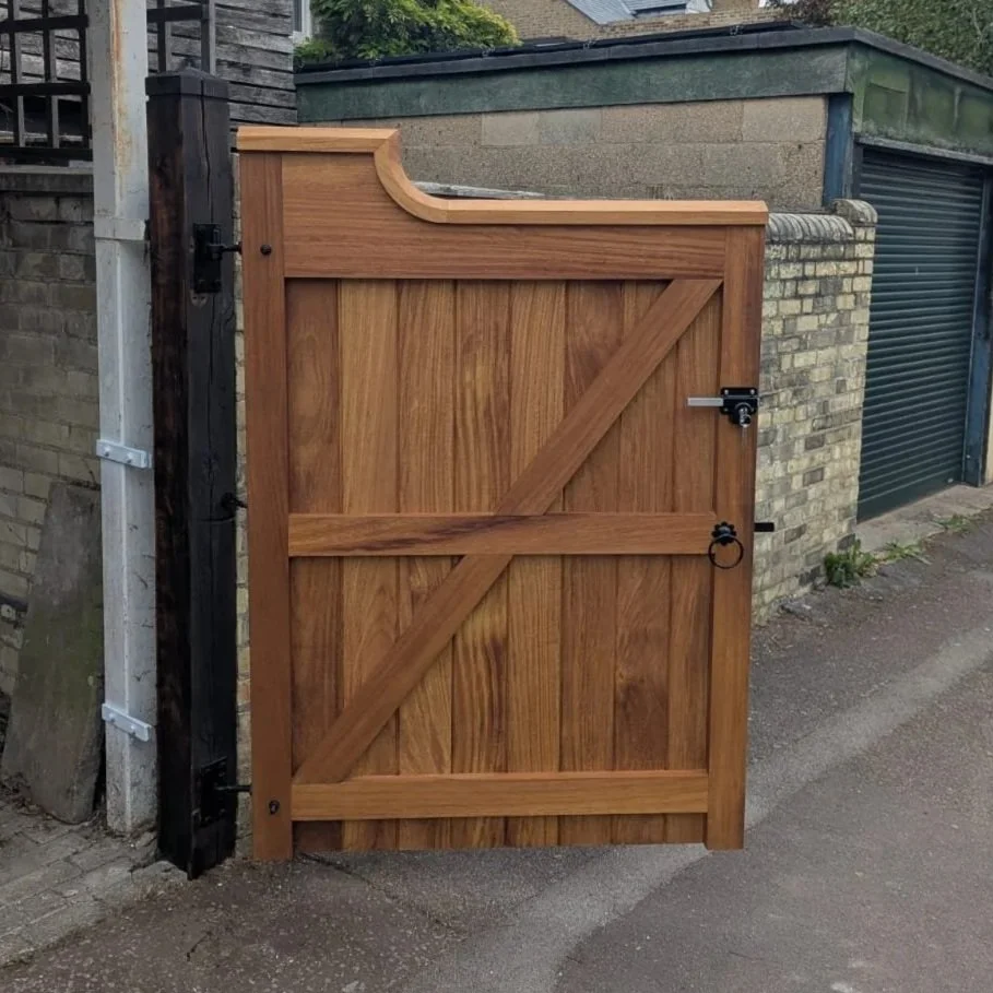 A tall wooden gate with a curved top on the left side, secured with a latch, attached to a brick wall and a black post. The gate is made of vertical wooden planks with diagonal and horizontal supports, and the setting appears to be an alley or backyard entrance.