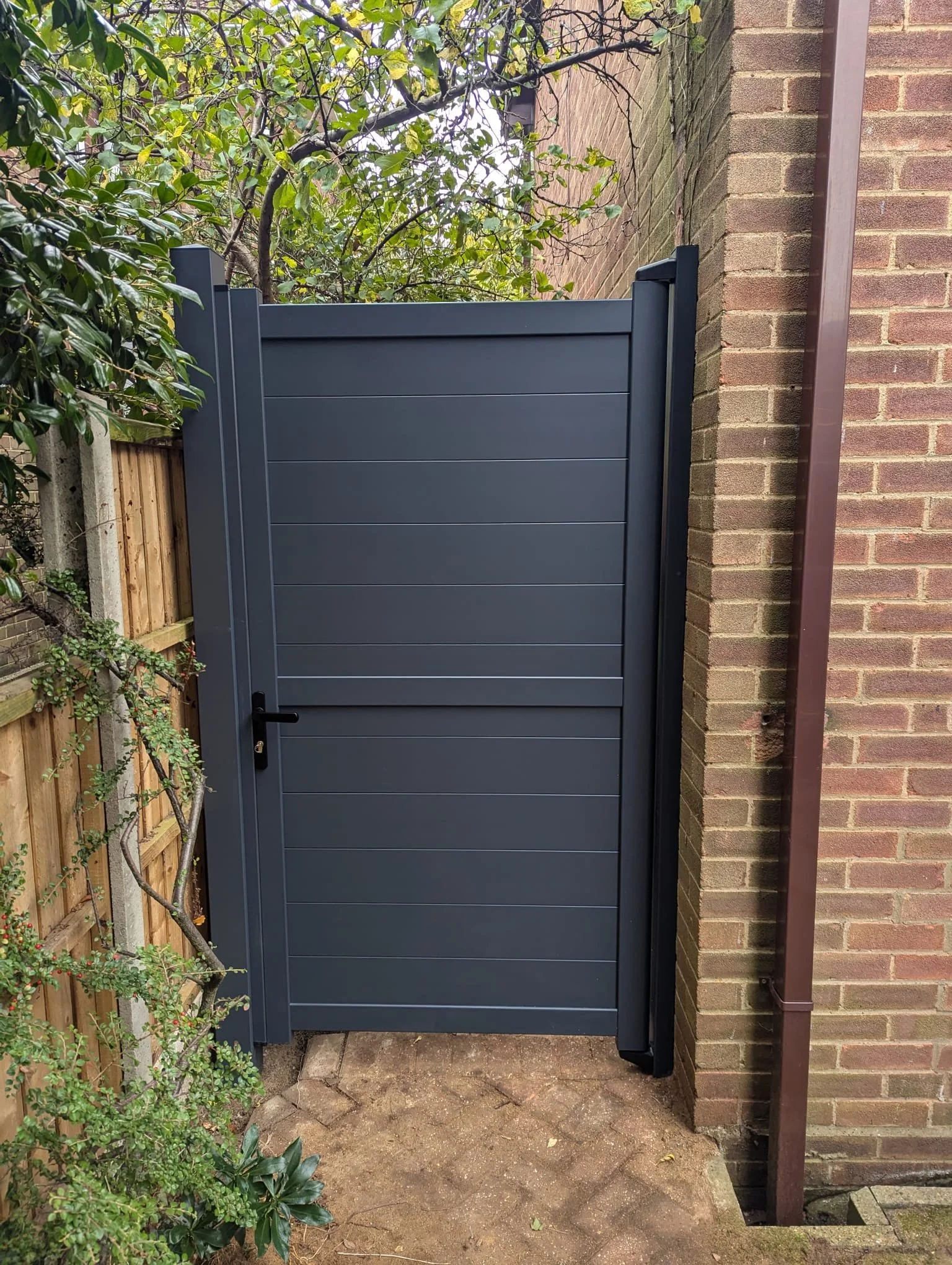 A dark gray metal gate with horizontal panels, attached to a brick wall on the right, leading to an outdoor space with a wooden fence and greenery in the background.