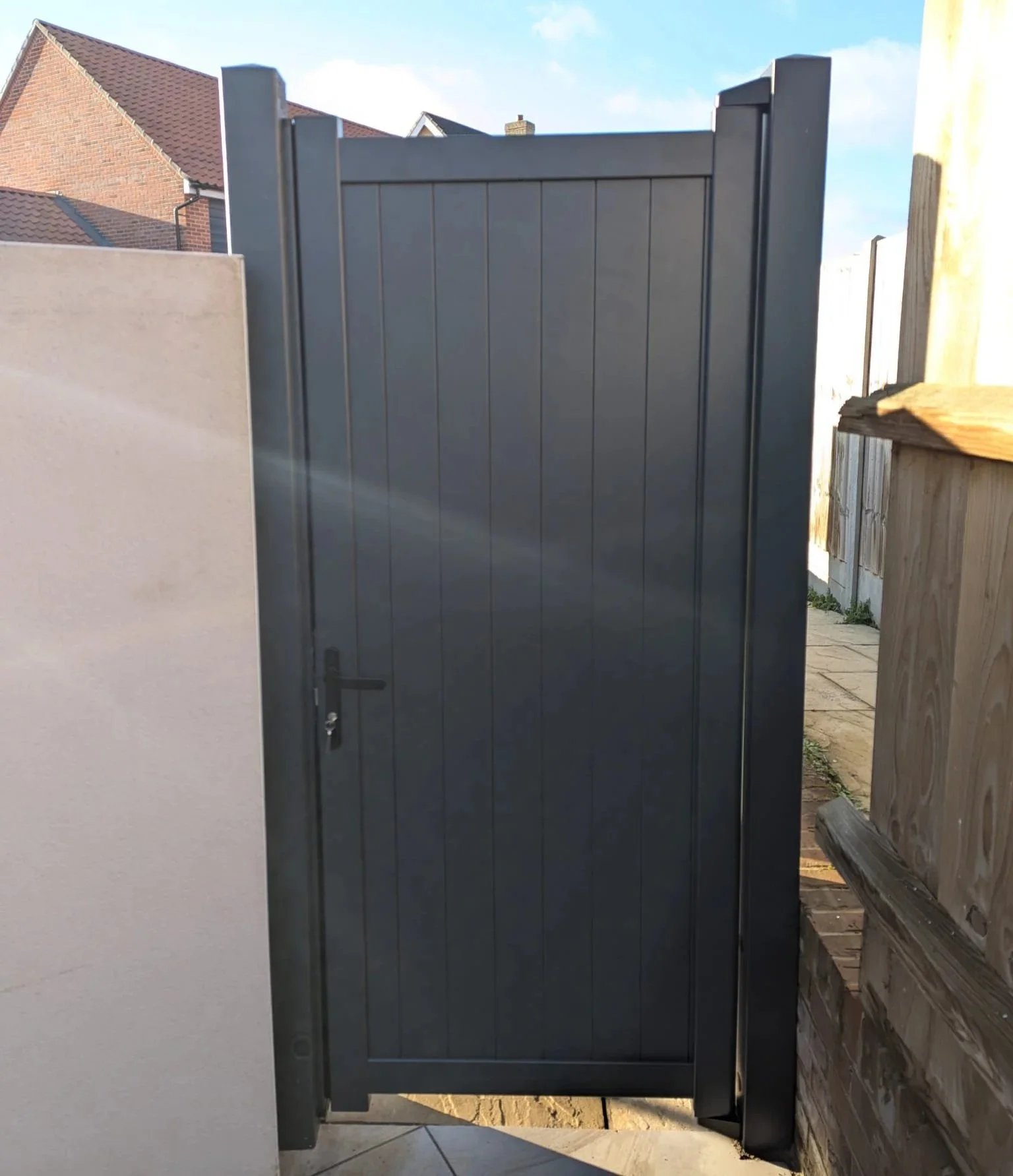 A dark gray metal gate with vertical panels, partially open, with a black handle on the left side, set between a beige wall on the left and a wooden fence on the right, with houses and a blue sky in the background.