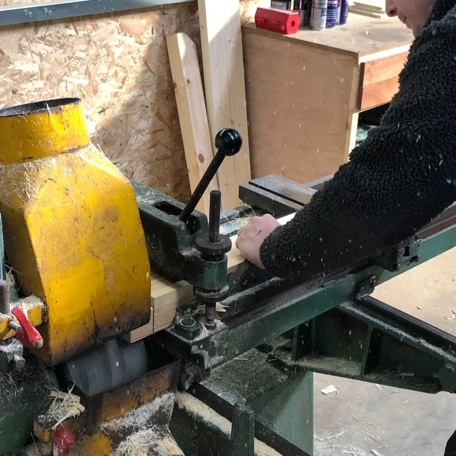 A person operating a woodworking machine in a workshop, visible with a black fleece sweater, working on a piece of wood.