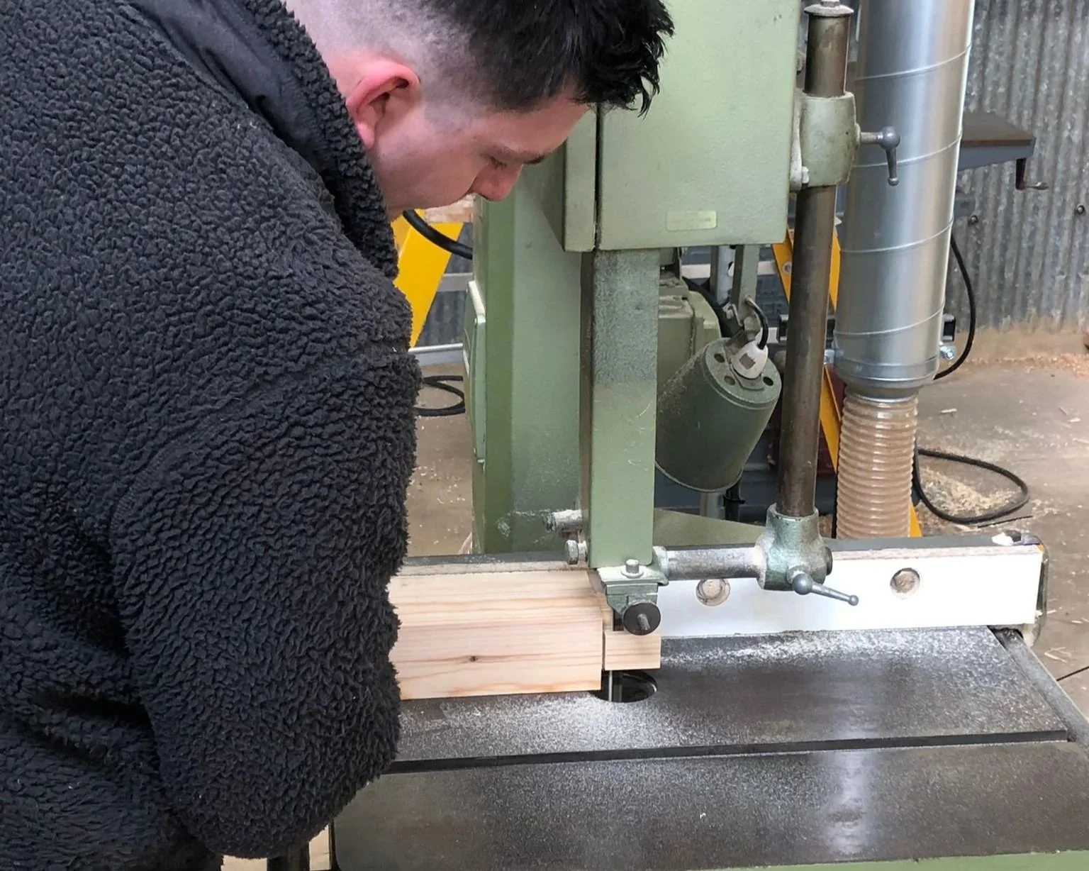 A person operating a green woodworking drill press machine, drilling into a piece of wood in a workshop.
