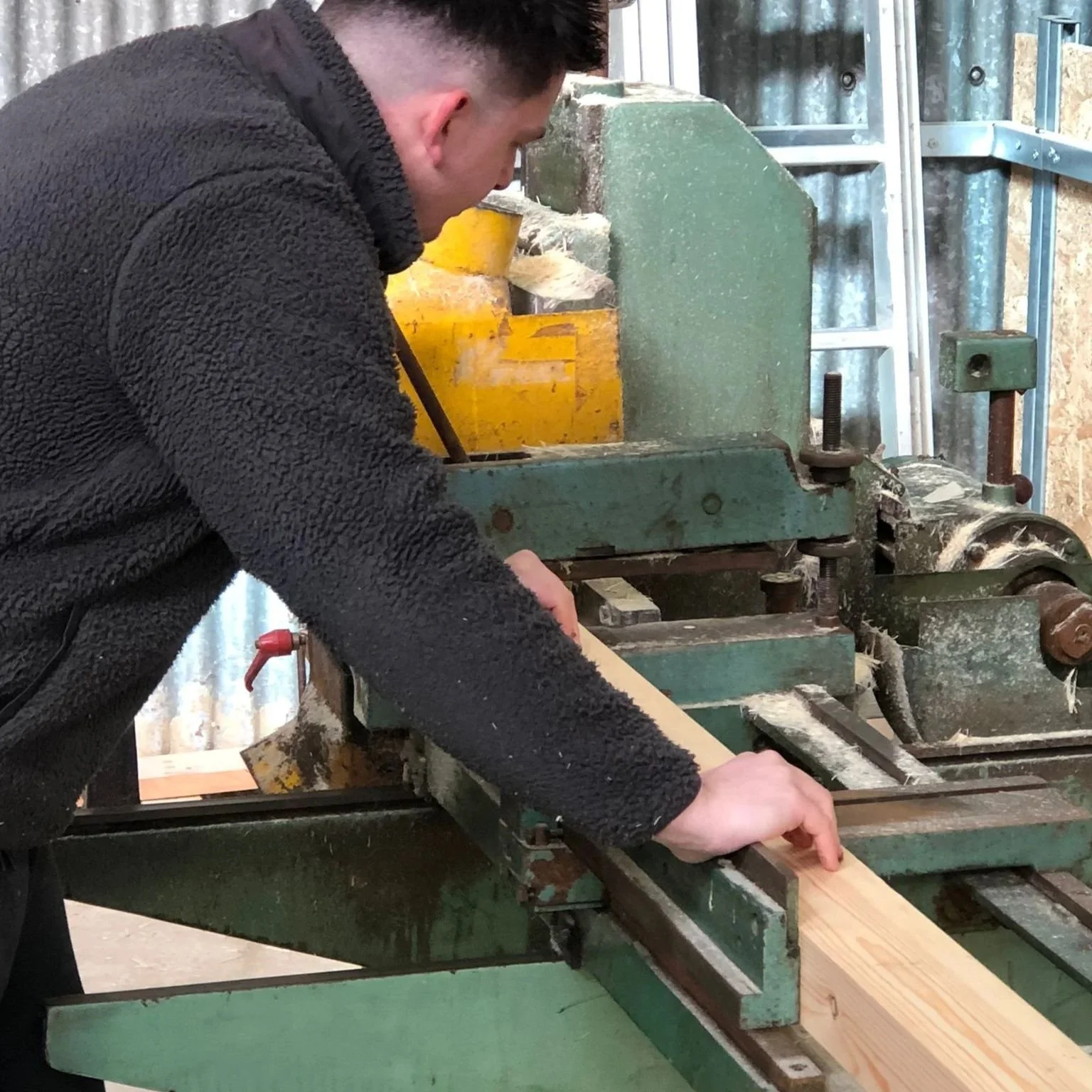 A man operating a green woodworking machine, cutting a wooden plank in a workshop with various tools and metal shelving in the background.