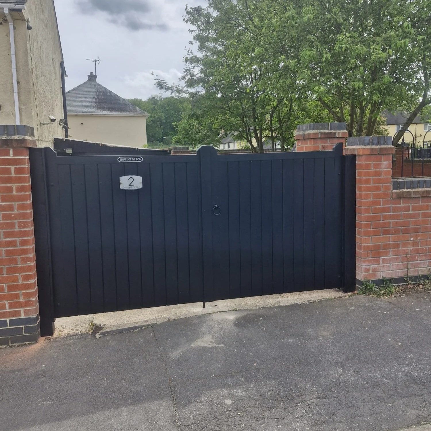 A black metal gate with a ring handle, flanked by brick pillars, opens to a driveway and leads to houses with trees in the background.