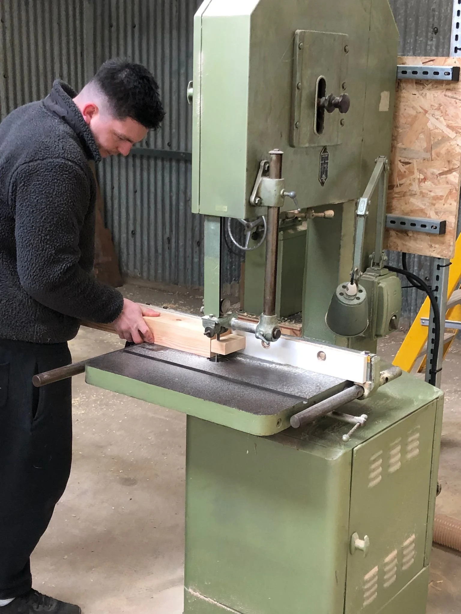 A man operating a large green bandsaw machine in a workshop, cutting a piece of wood.