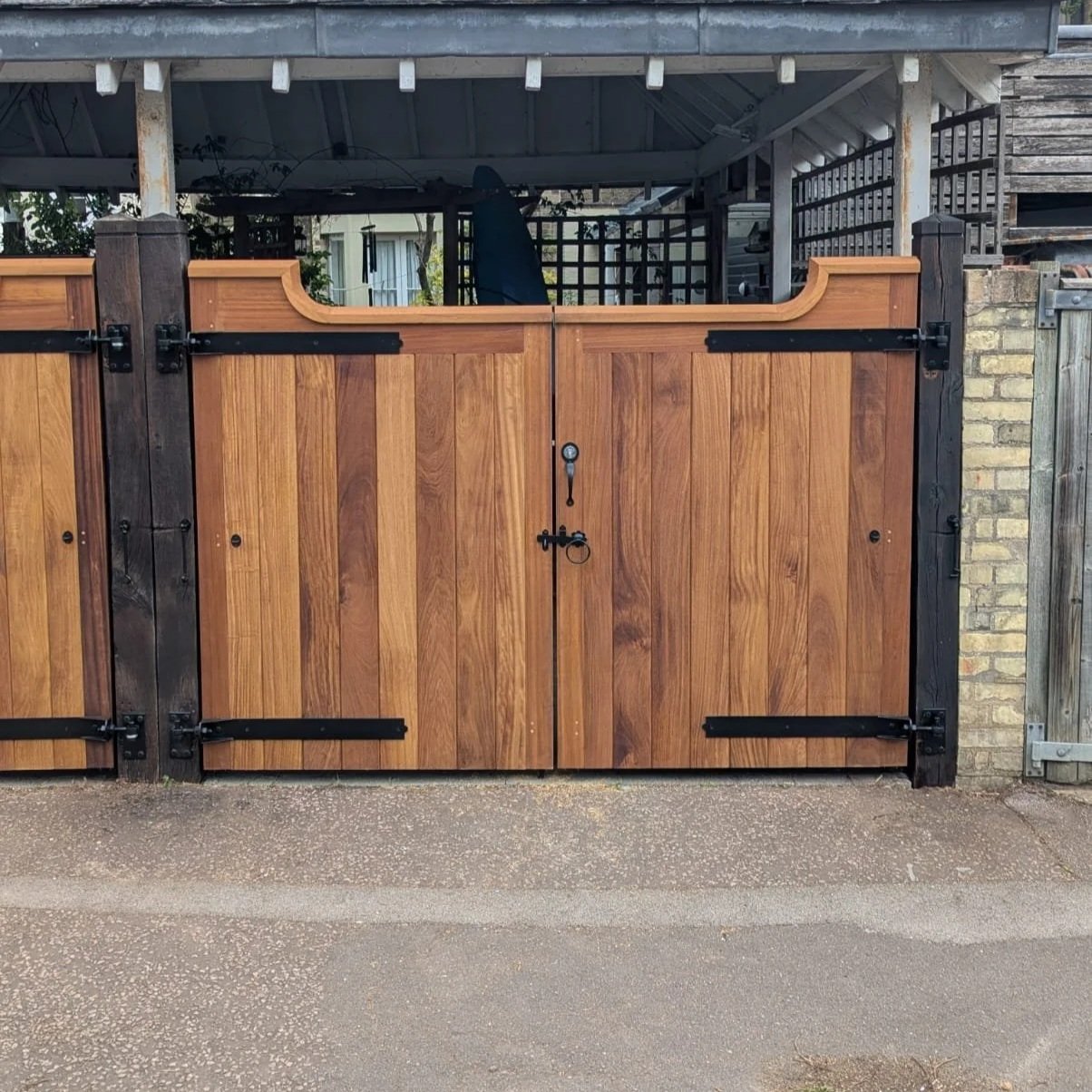A wooden gate with black hardware and a latch, part of a fenced yard, with a covered patio and a tree in the background.