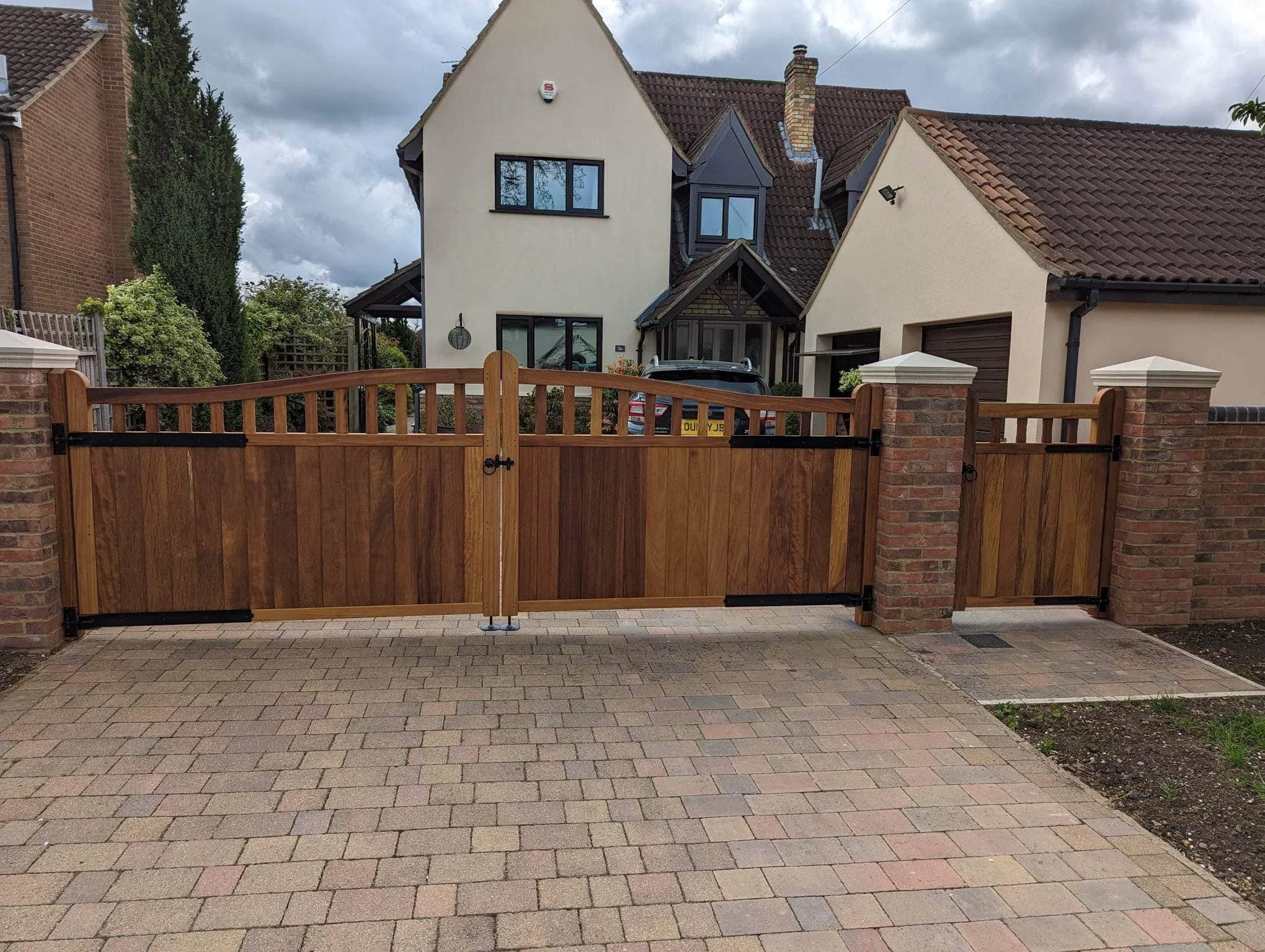 A wooden driveway gate with black hinges and latch, set in brick pillars, opening to a paved driveway leading to a house with beige walls, reddish-brown roof tiles, and multiple windows. There are cloudy skies overhead.