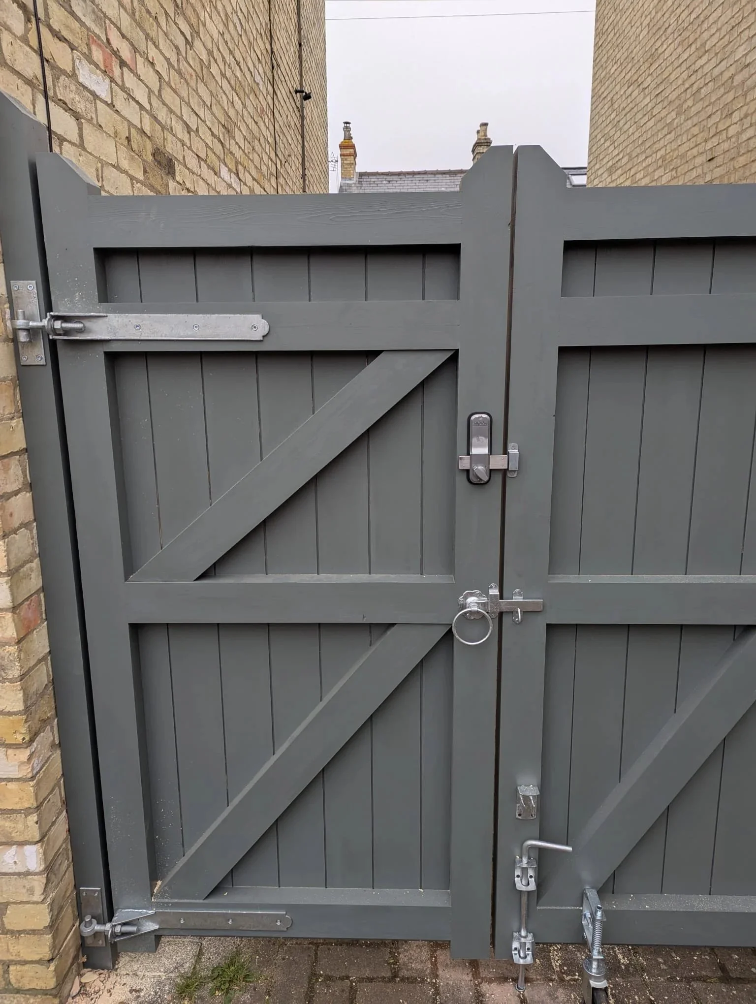 A gray wooden gate with metal hardware, visible lock, latch, hinges, and a ring handle, attached to brick walls on each side, with a paved ground in front.