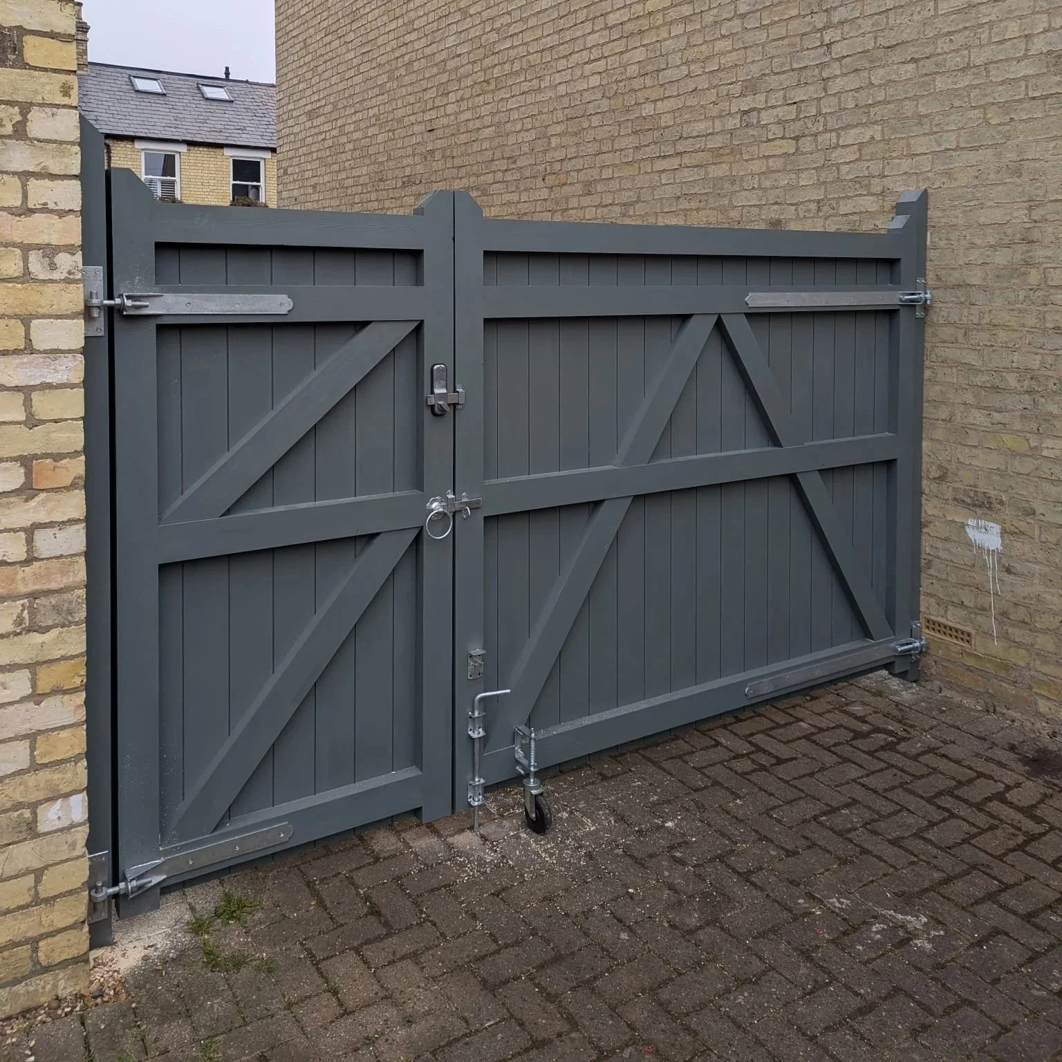 Gray double gates made of wood and metal, secured with locks, situated between brick walls on a paved ground.