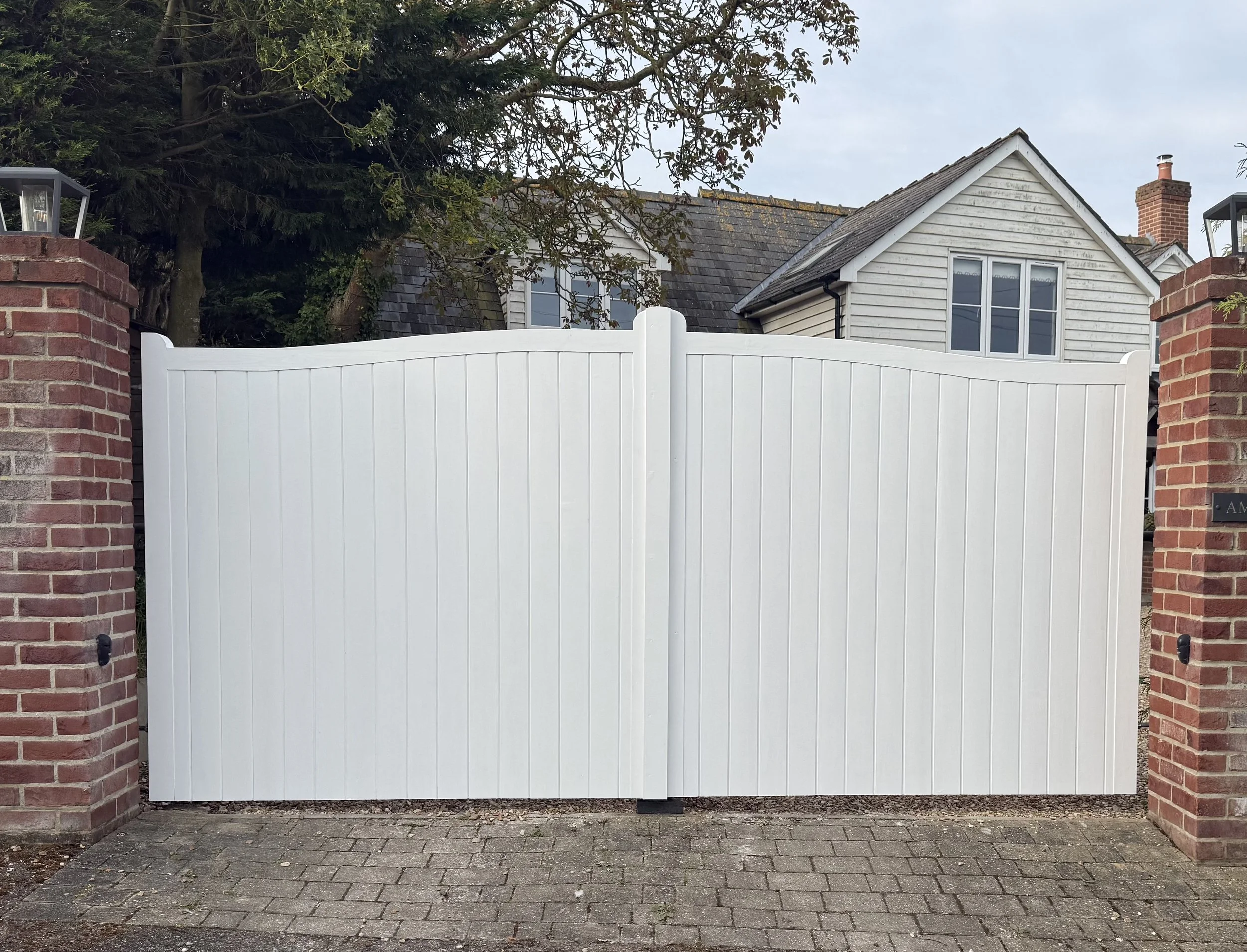 A white privacy fence with two brick pillars on either side, located on a brick sidewalk. Behind the fence, there's a house with a gray roof, white siding, and a brick chimney. The house has two windows, and there are trees and shrubs around it.
