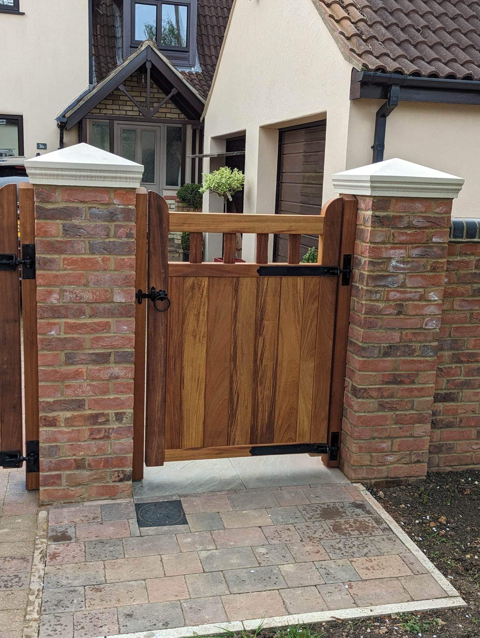 A wooden gate attached to brick columns in front of a house with a tiled roof and a driveway.