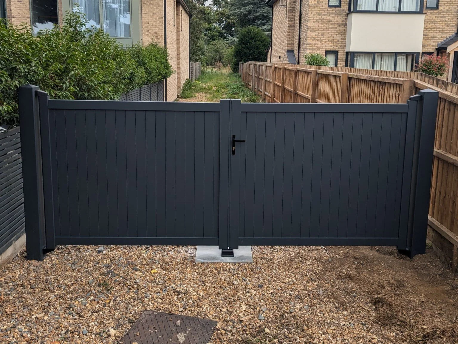 Closed black metal gate with vertical slats at the entrance of a residential property, surrounded by gravel ground and fencing on both sides.