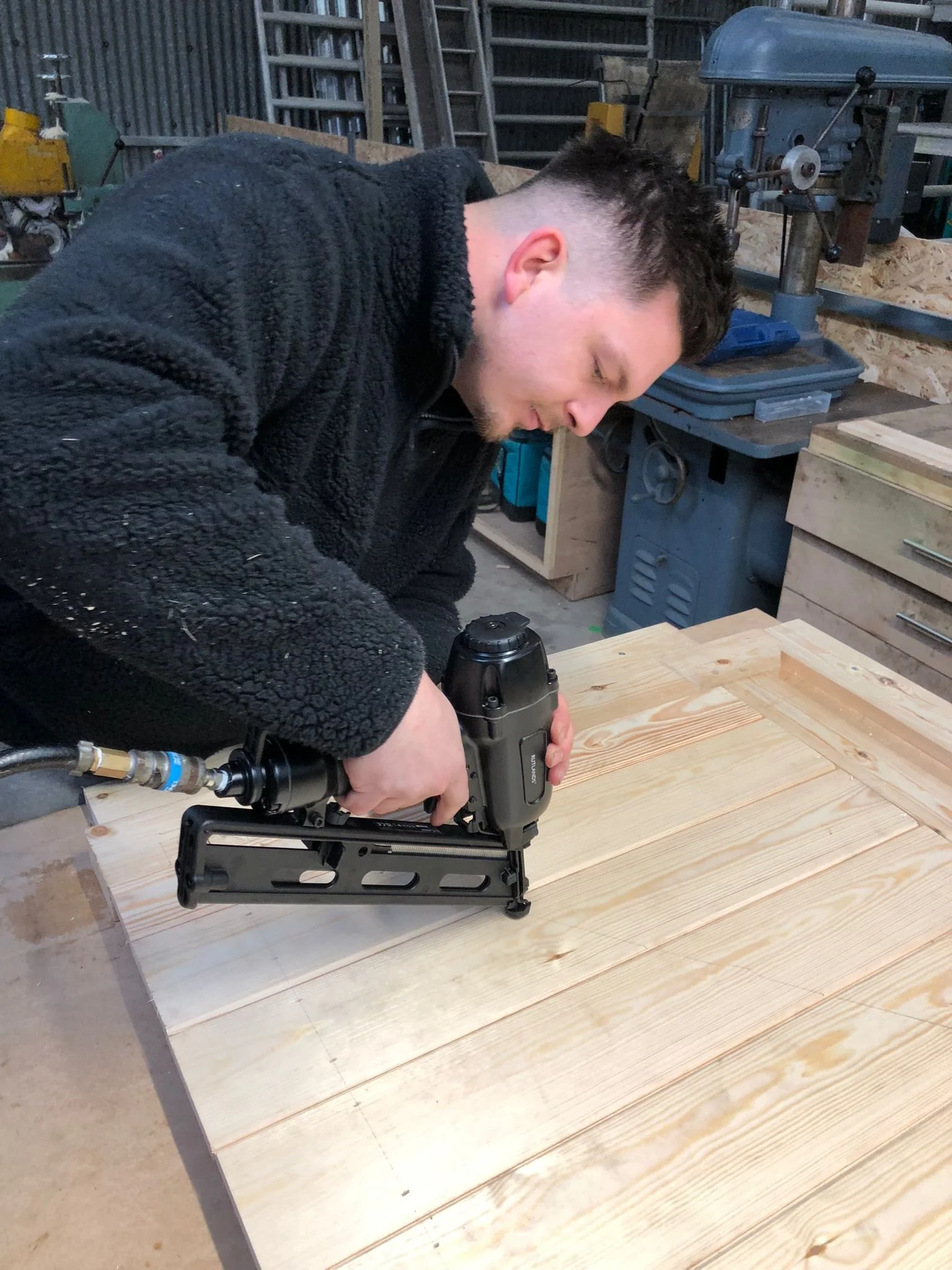 Man using a nail gun on a wooden workbench in a woodworking shop.
