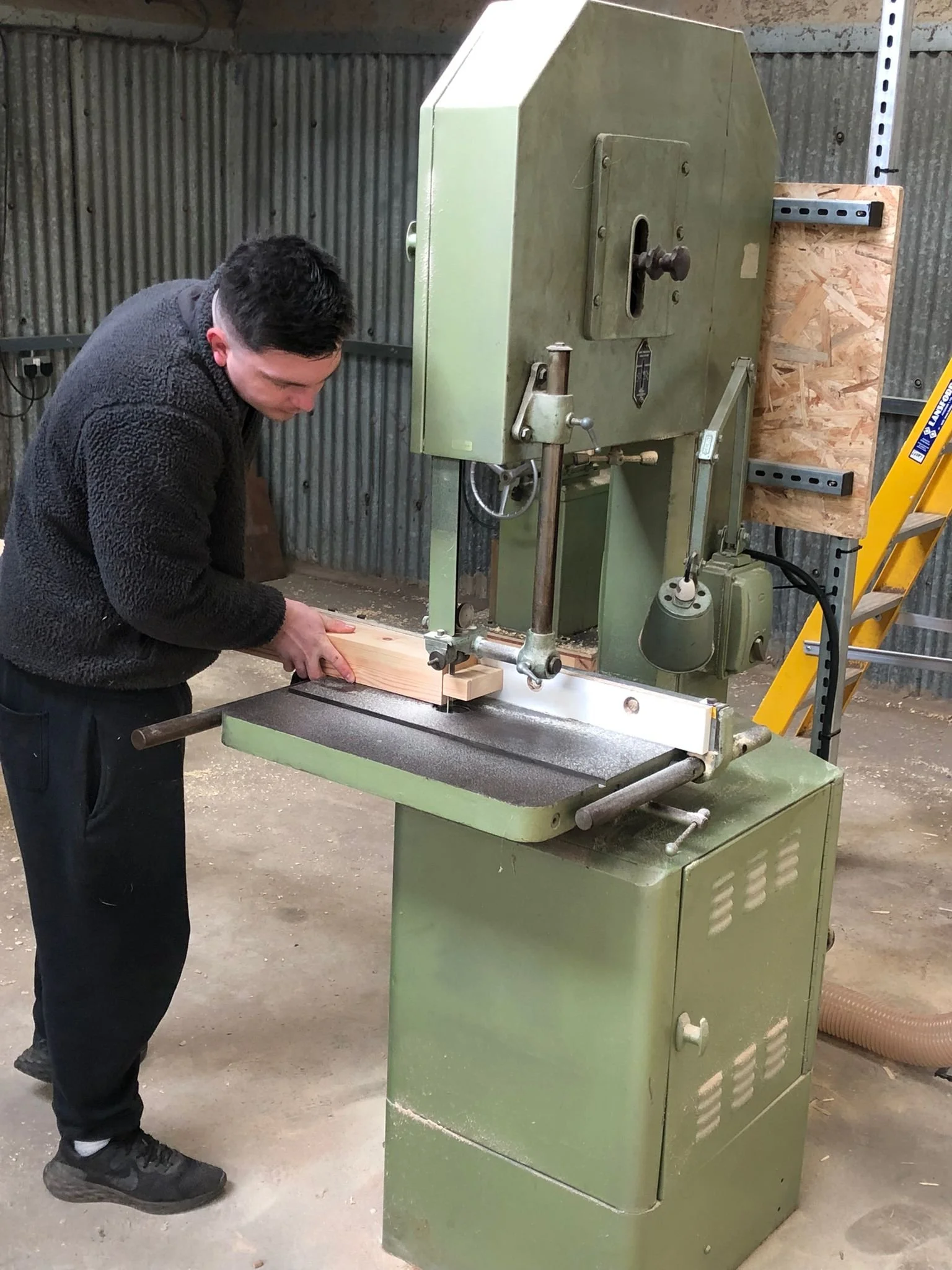 A person operating a green bandsaw to cut a piece of wood inside a workshop.