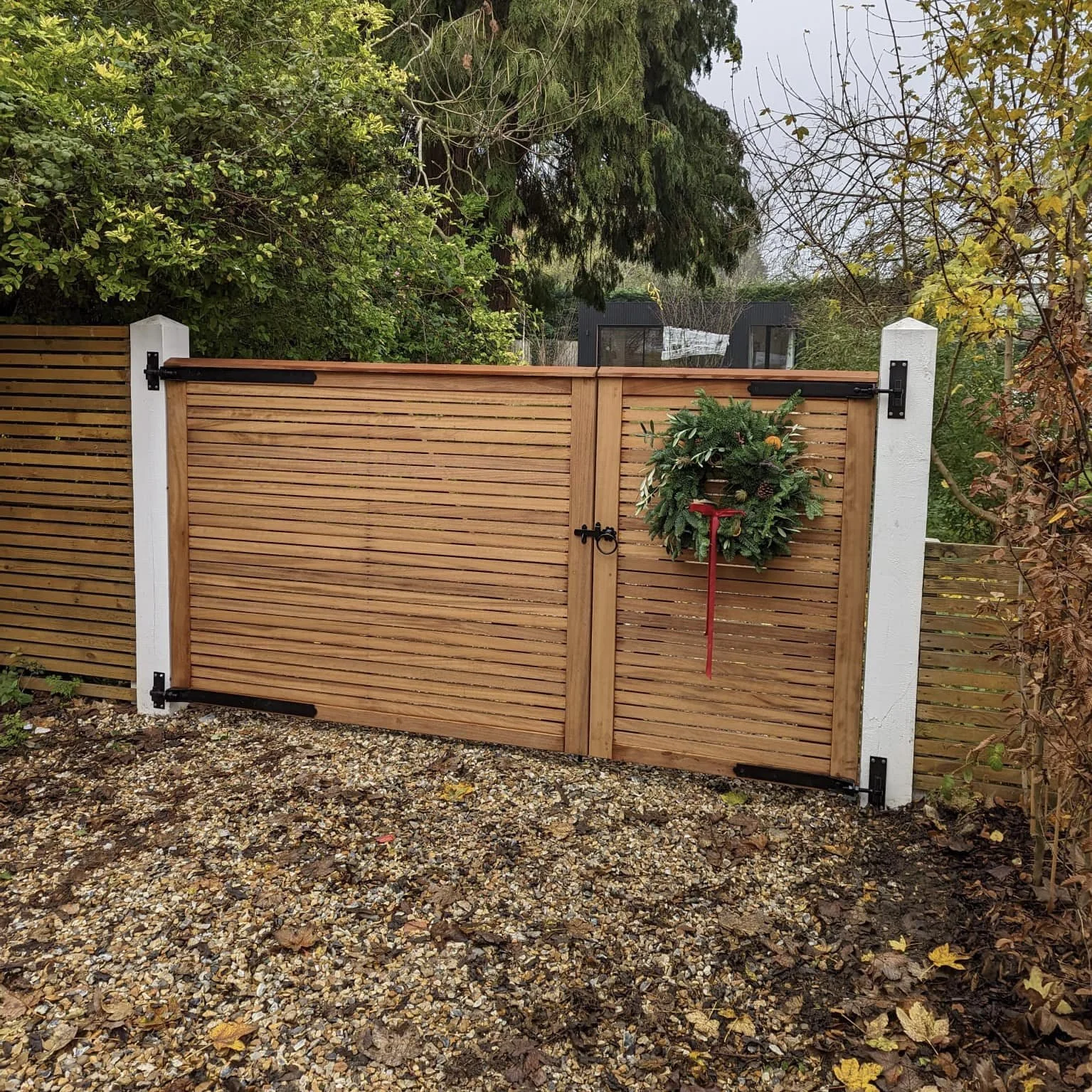 A wooden gate decorated with a Christmas wreath, situated between two white posts in a garden with trees and bushes.