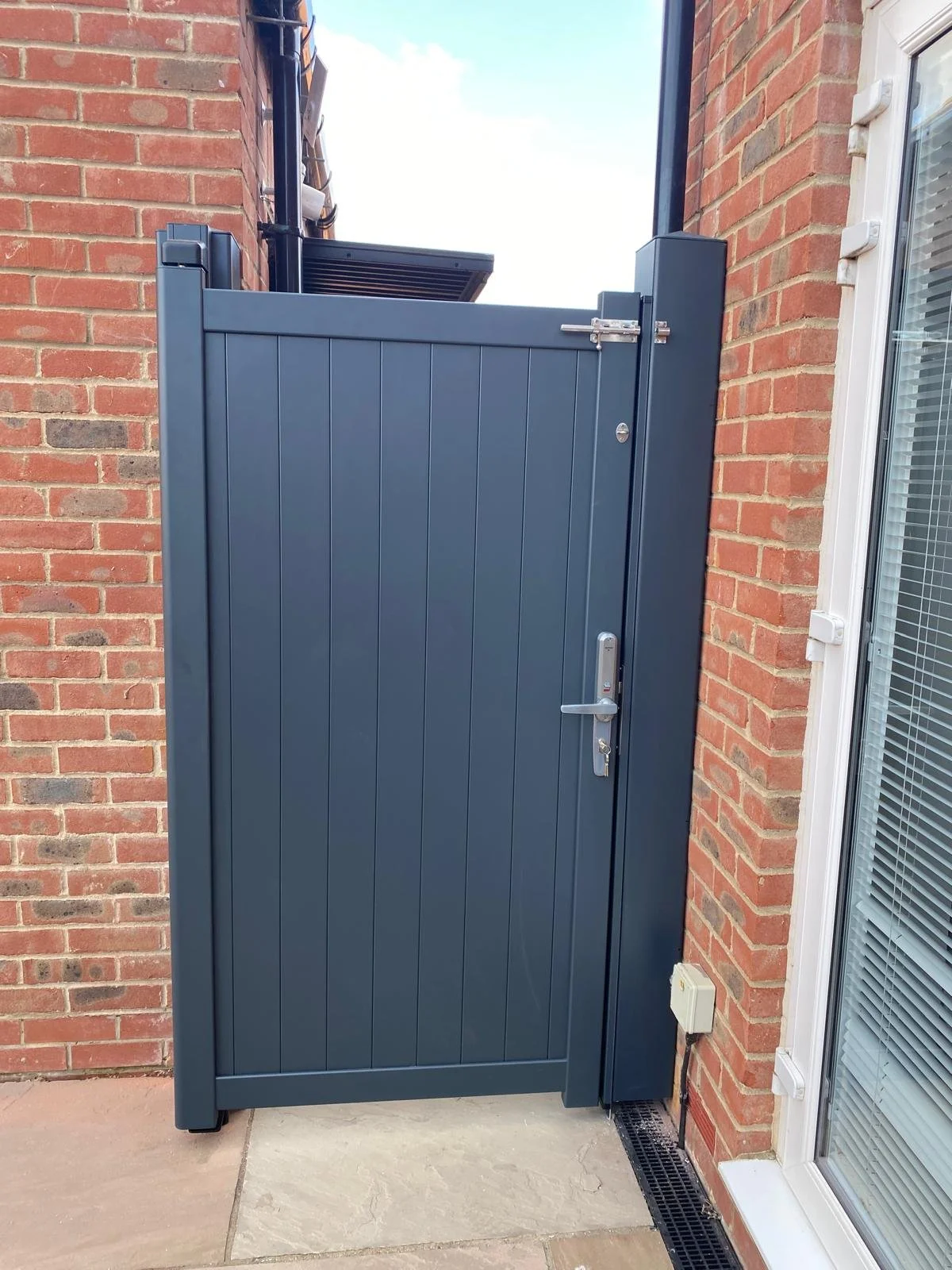 A dark gray exterior gate installed between a brick wall and a white-framed window, with a silver handle and lock, and a small white electrical box below.
