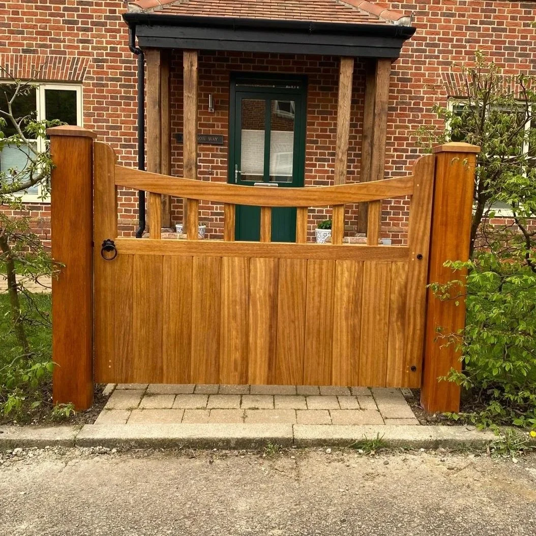 A backyard with a wooden gate, a gravel pathway, a garden bed, a lush green lawn, and a house with many windows and a brick chimney, surrounded by trees and bushes.