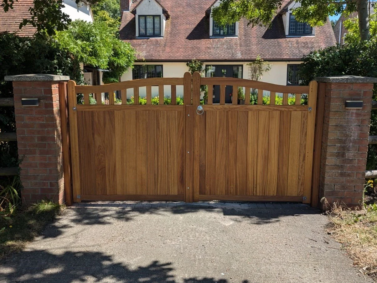 A wooden gate with a curved top, set between two brick pillars, blocking a driveway in front of a house with a red-tiled roof and multiple windows, surrounded by greenery.