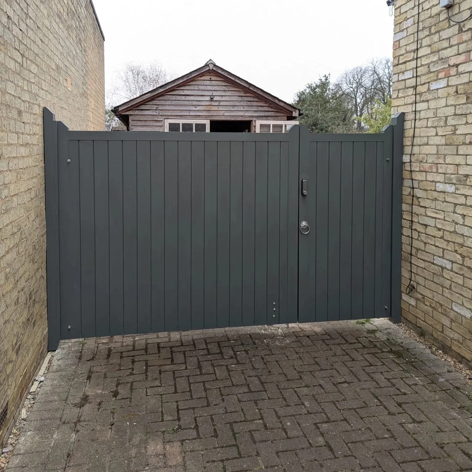 Wooden gate with brick pillars in front of a house with a red-tile roof and dormer windows, surrounded by greenery and trees.