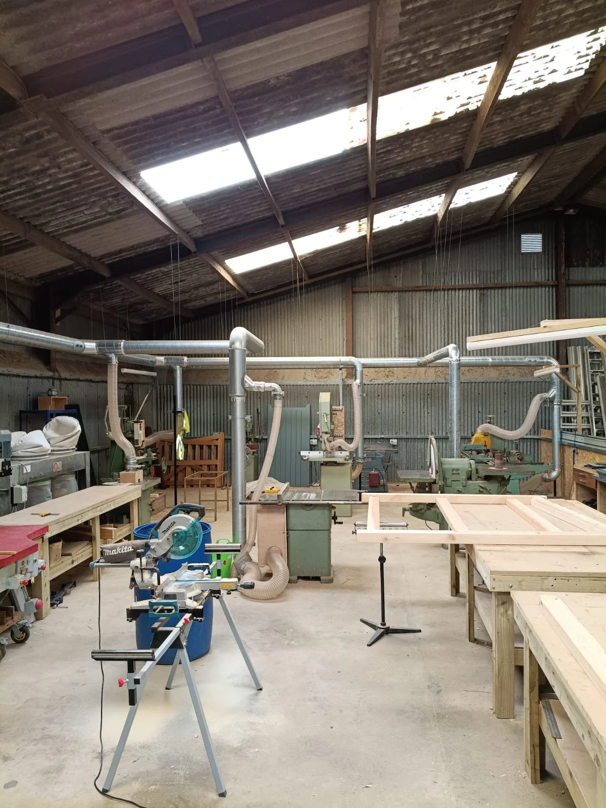 A woodworking workshop with various power tools, wooden workbenches, and ventilation ducts, inside a spacious industrial building with a high, corrugated roof.