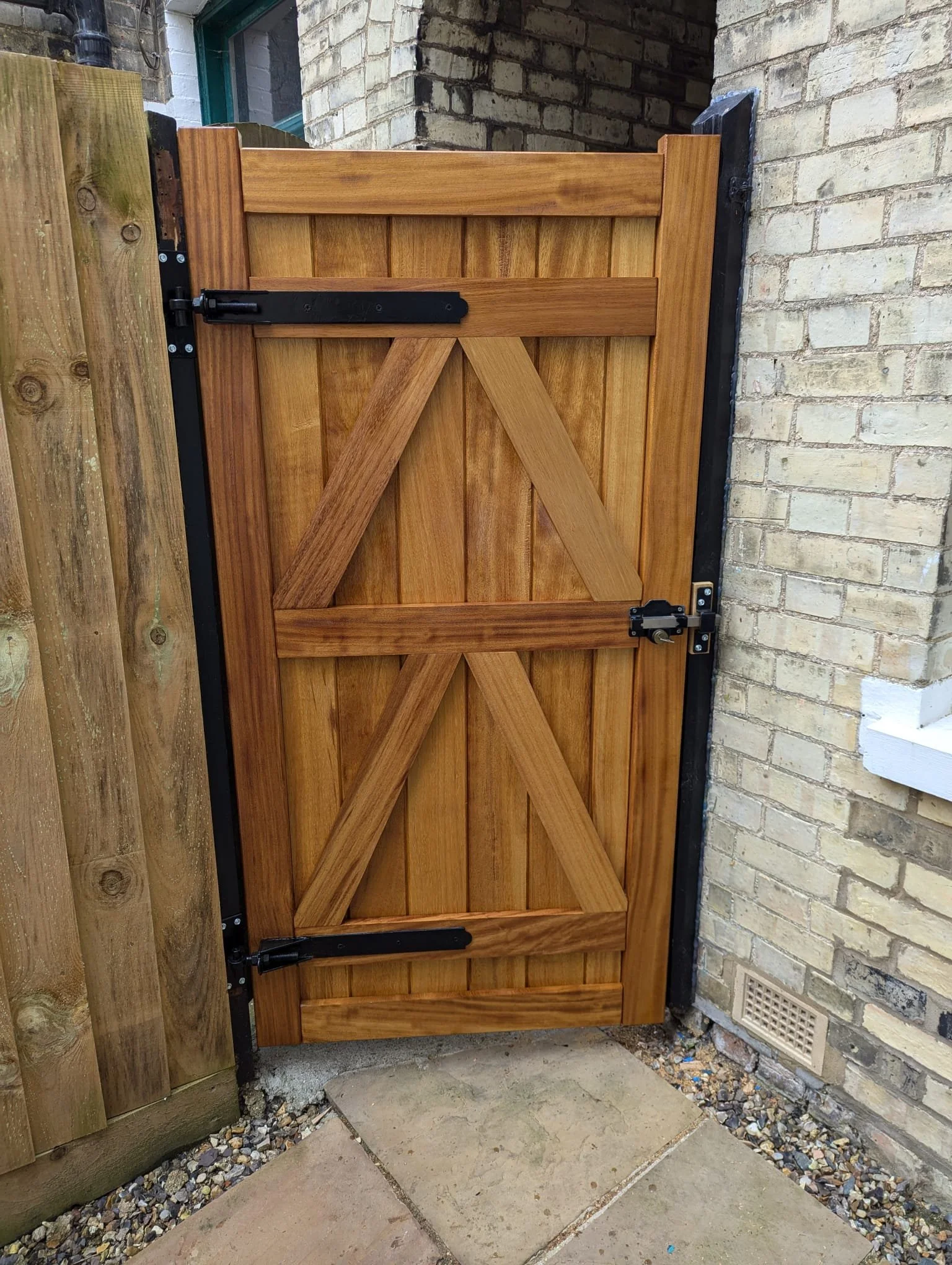 A wooden garden gate with black metal hinges and latch, installed between a brick wall and wooden fence.
